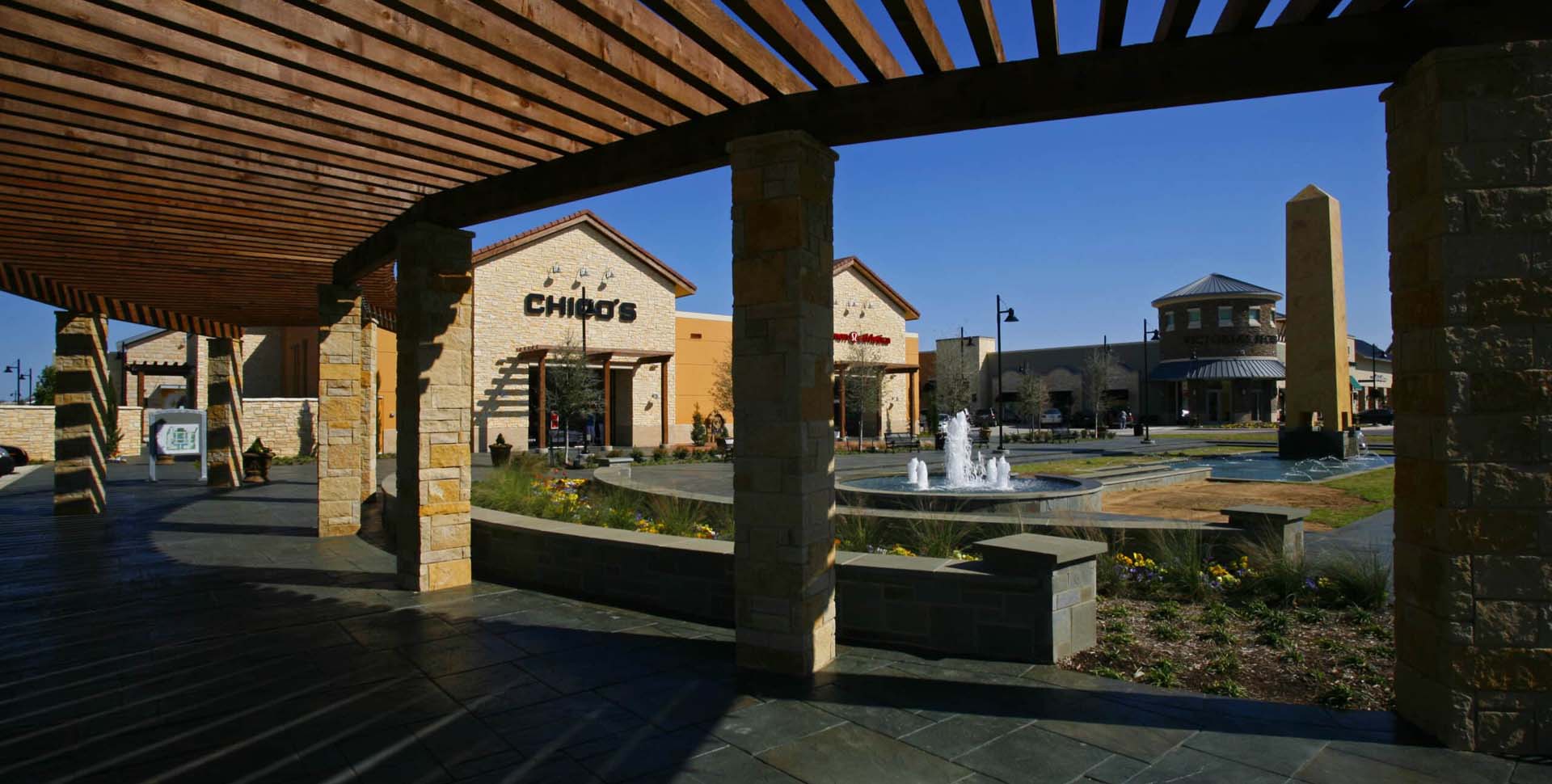Outdoor shopping center with stone columns, a water fountain, landscaped areas, and storefronts including Chico’s under a clear blue sky.