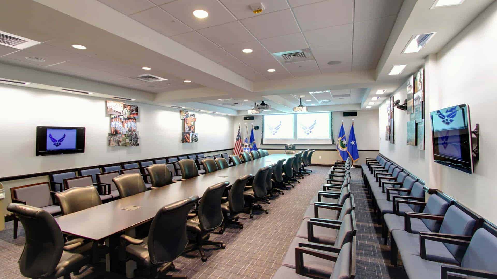 Modern conference room with long tables, office chairs, side seating, wall-mounted screens displaying a military insignia, and flags positioned at the front of the room.