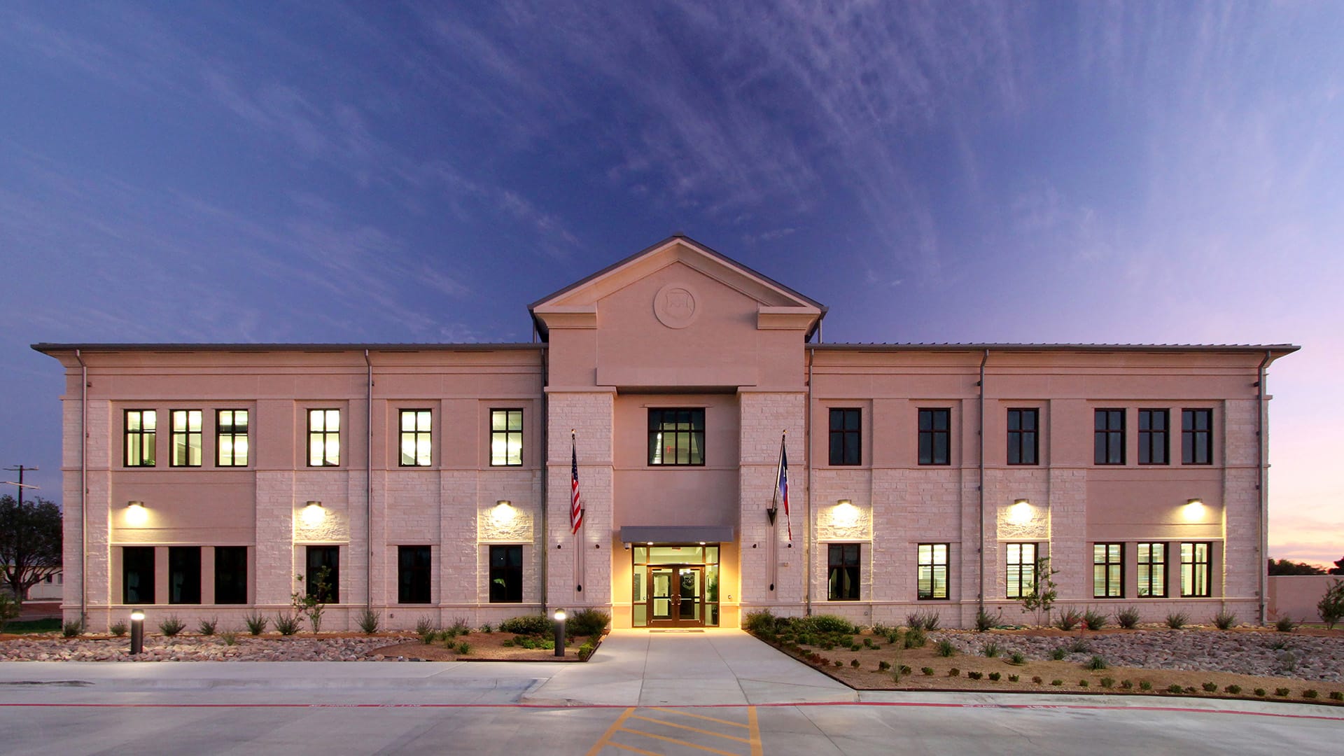A two-story beige office building with large windows, American and state flags at the entrance, and exterior lights on at dusk.