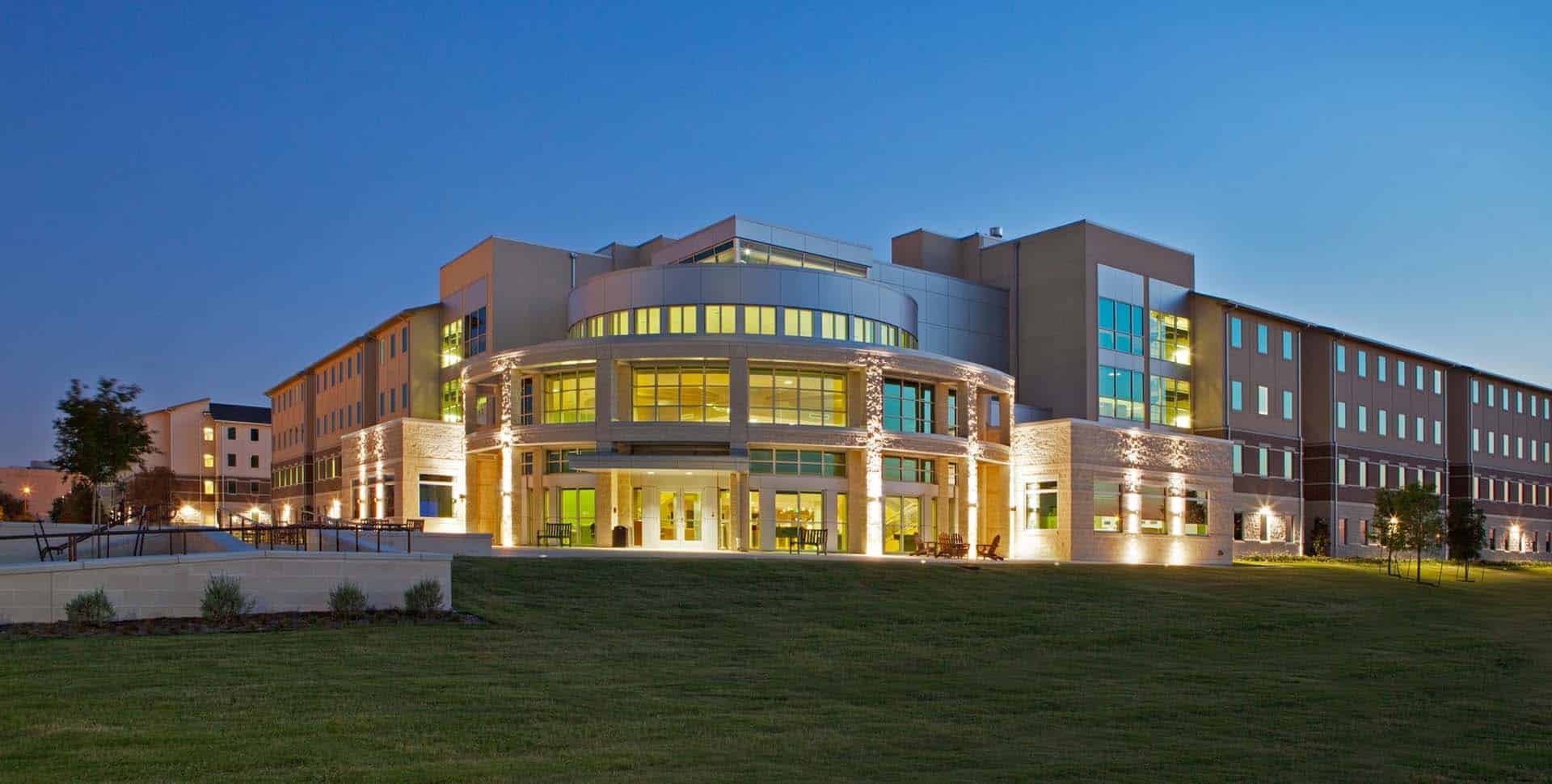 Modern, multi-story building with large glass windows and exterior lighting, set on a grassy lawn at dusk.
