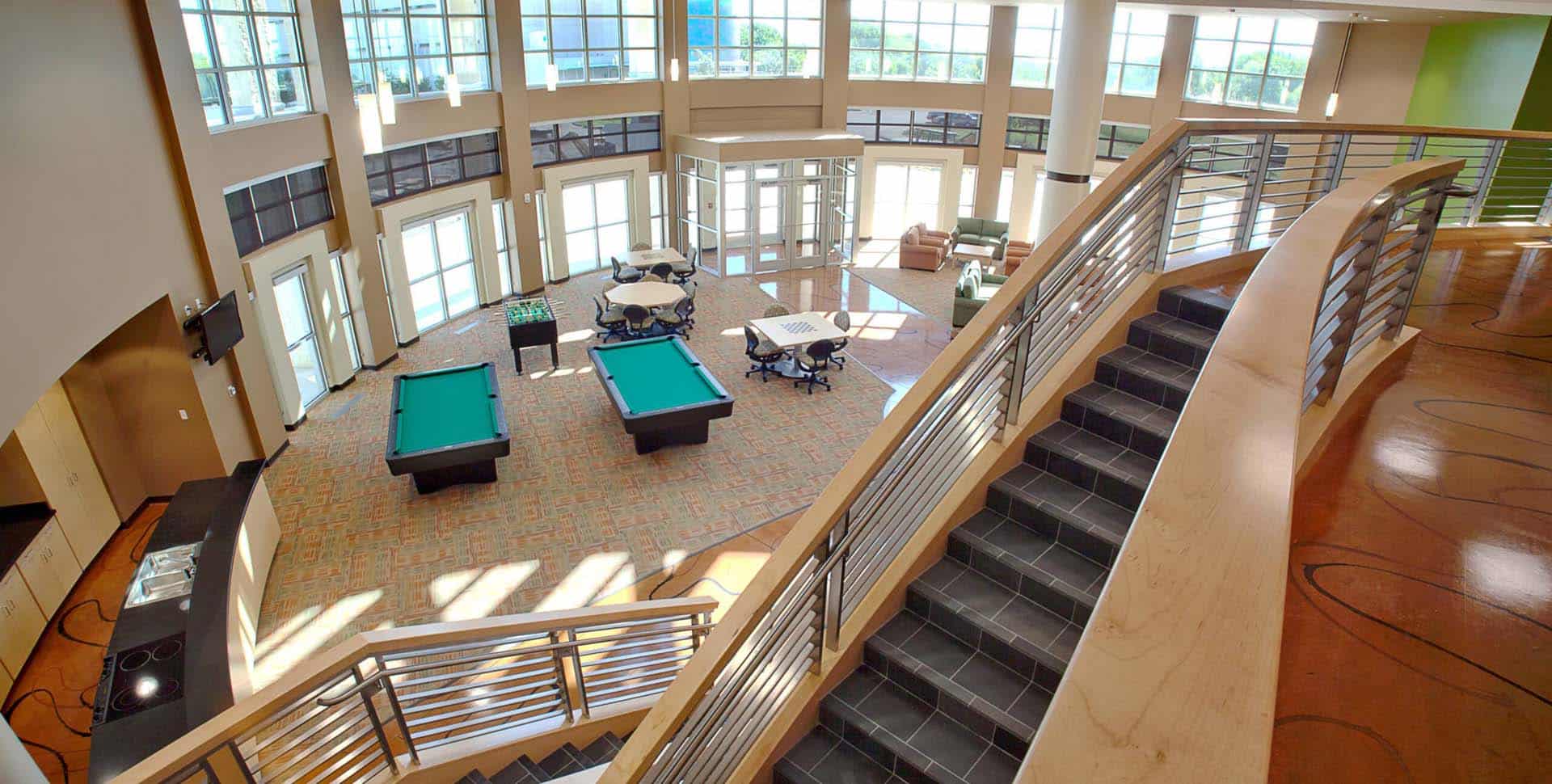 Spacious recreation room with large windows, pool tables, foosball, lounge chairs, tables, and a staircase with metal railings.