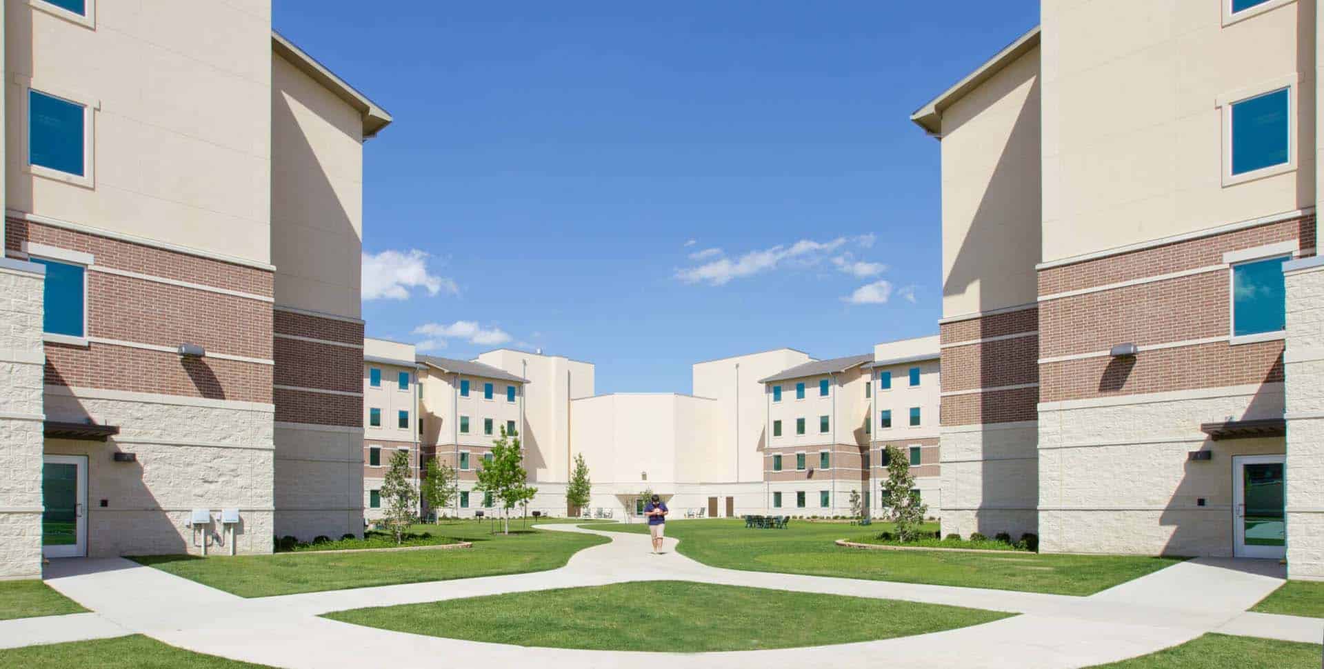 A person walks along a pathway between two four-story residential buildings with lawns, under a clear blue sky.