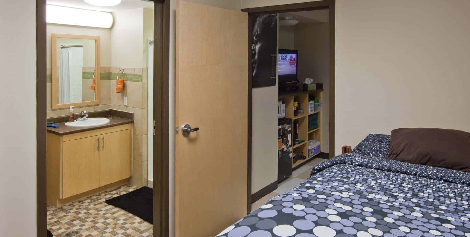A dorm room with a bed, open door to a bathroom with a sink and mirror, and shelves holding books and a TV.