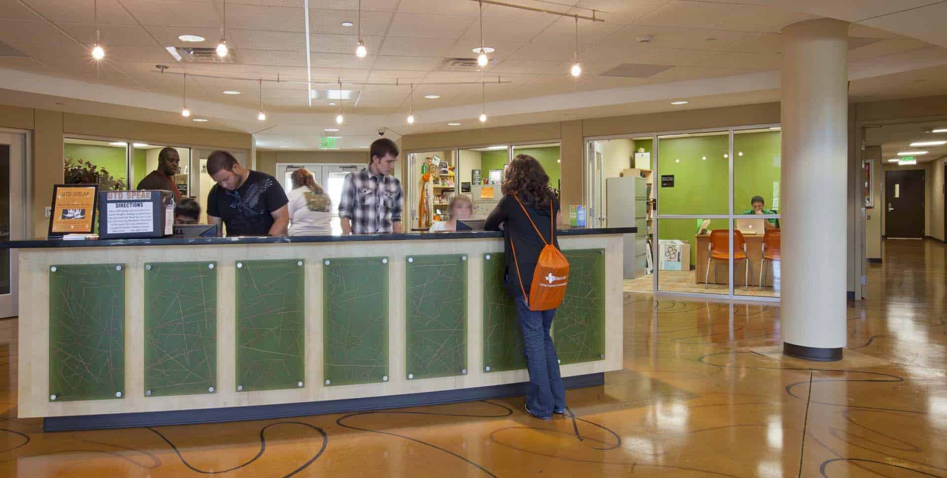 Several people stand at a reception desk in a modern, brightly lit office lobby with green accents and glass-walled rooms in the background.