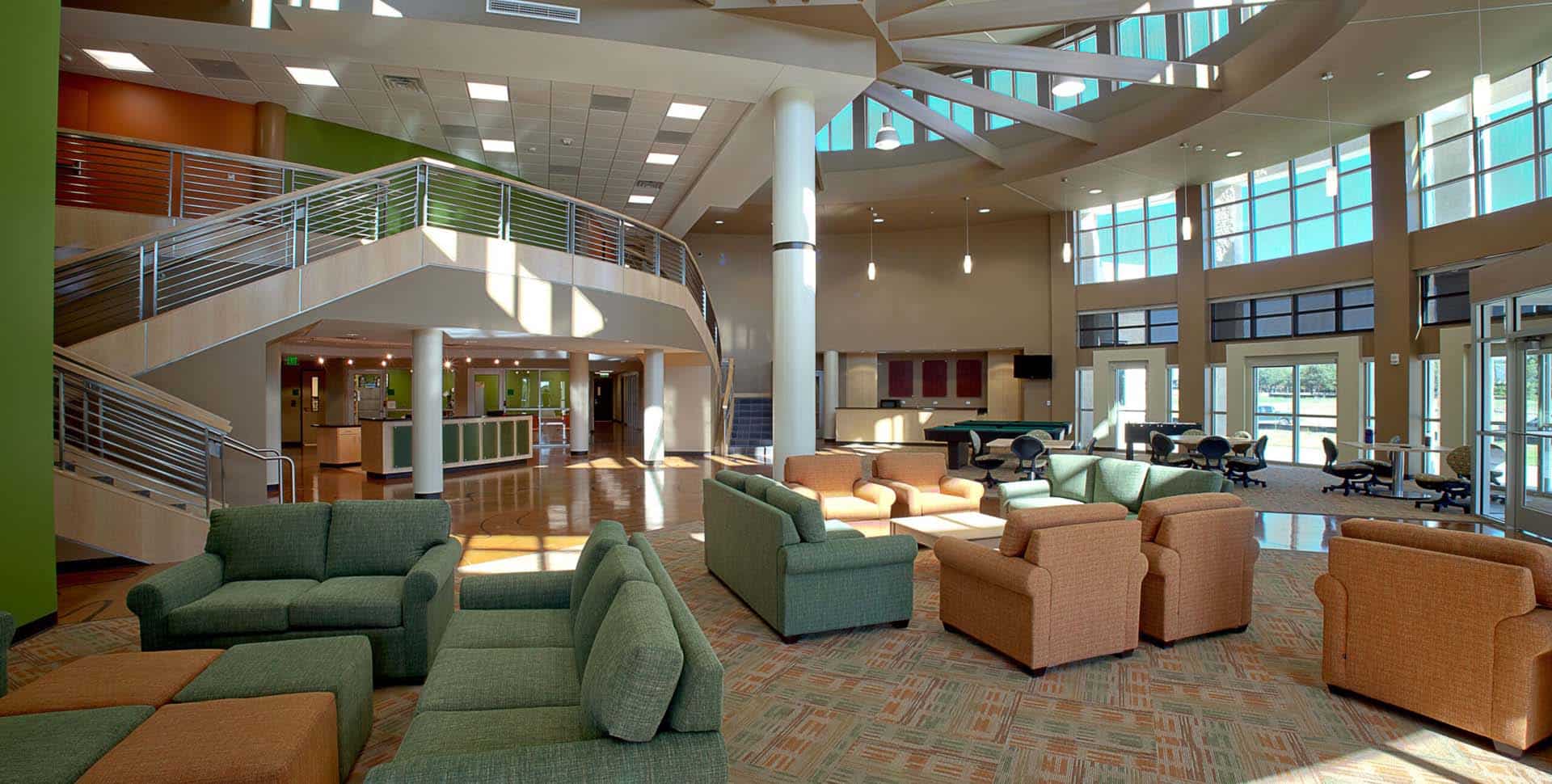 Modern lobby with green and beige armchairs, large windows, a staircase, and open seating area with natural light.
