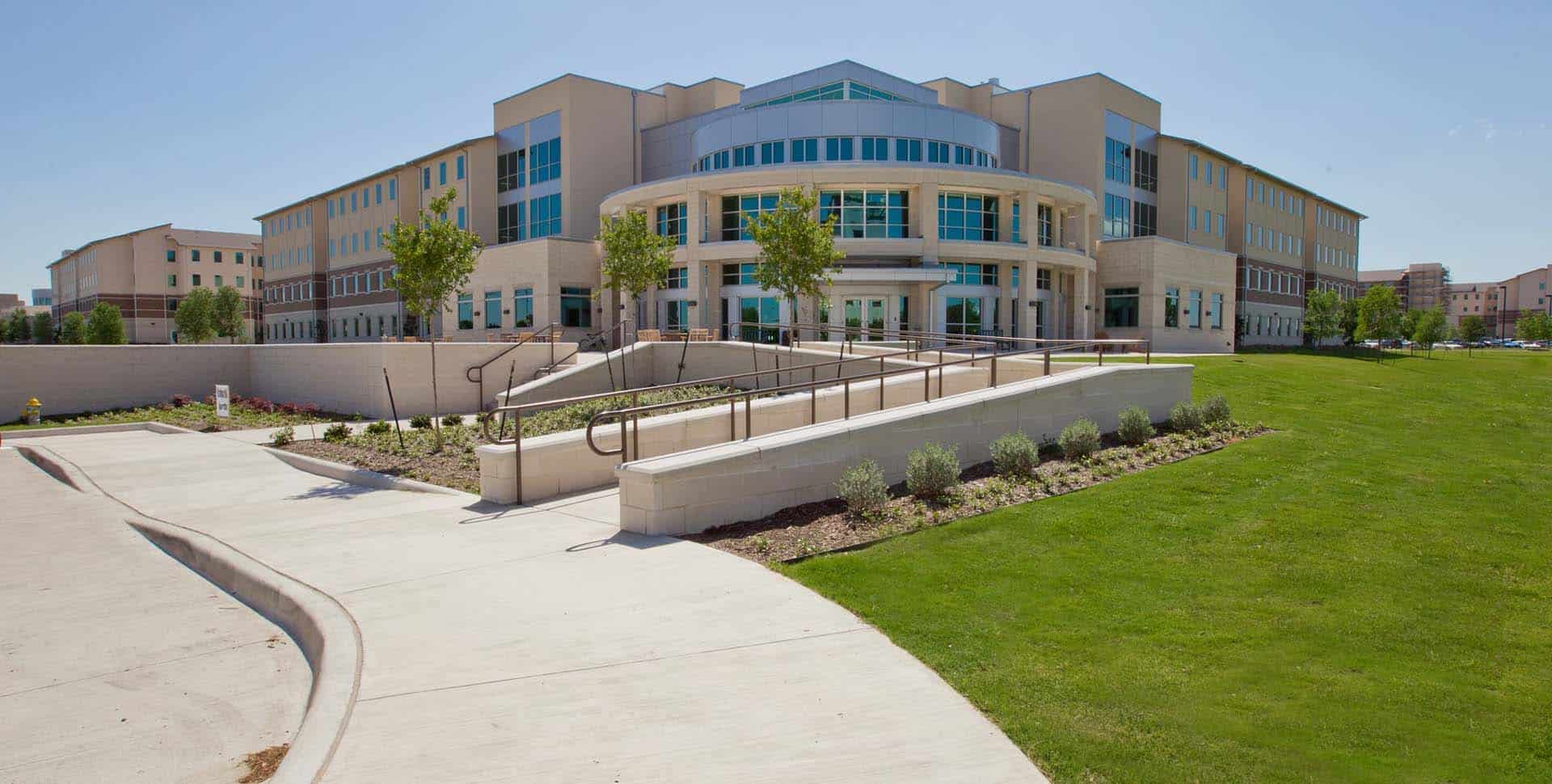 Modern multi-story building with large windows, surrounded by landscaped lawns and walkways, under a clear blue sky.