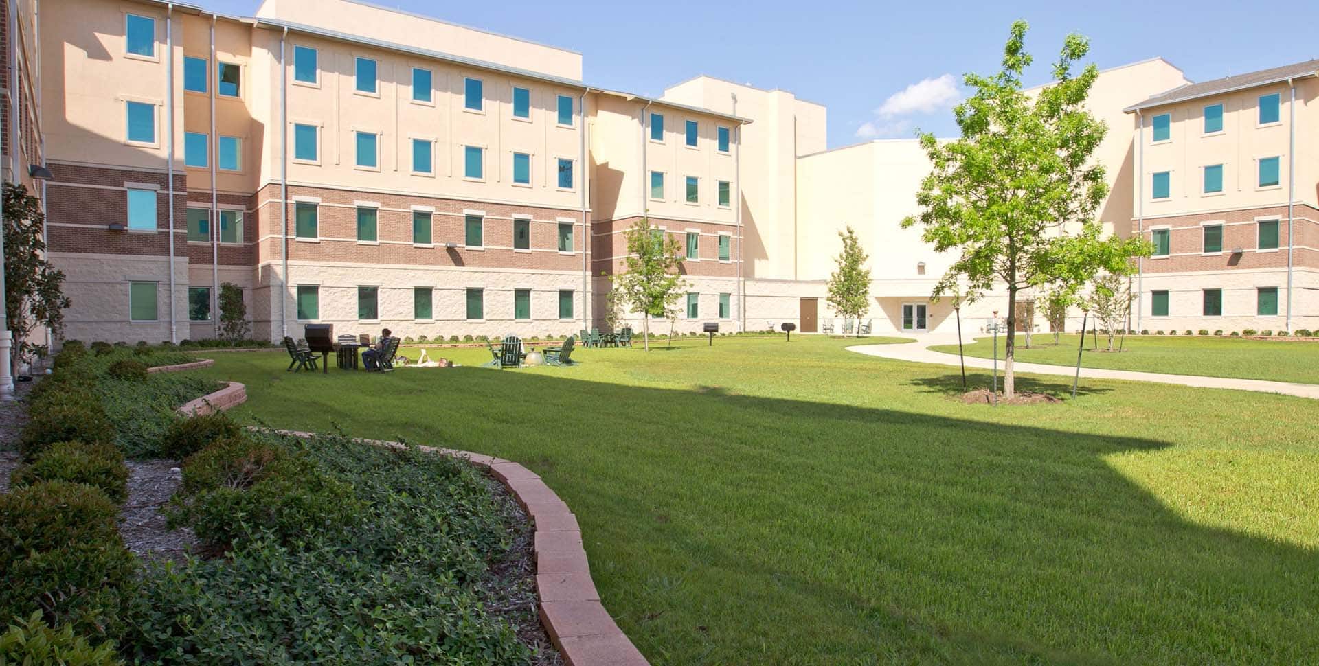 A three-story dormitory building surrounds a grassy courtyard with a few trees, bushes, benches, and outdoor seating under a clear sky.