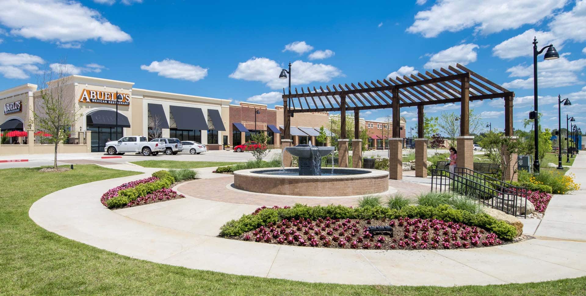A shopping center with a central fountain, landscaped flower beds, a pergola, and storefronts under a blue sky with scattered clouds.