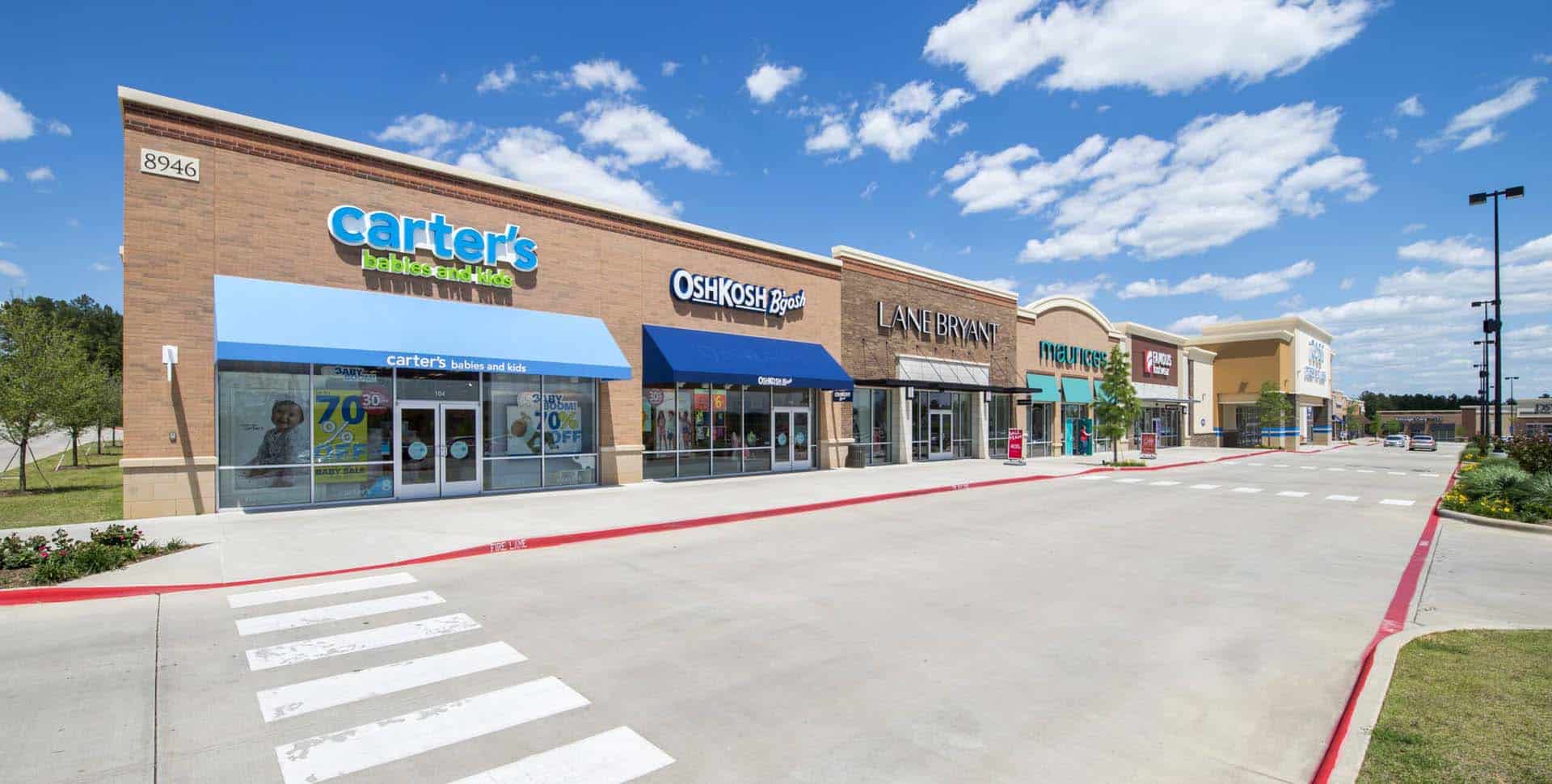 A row of retail stores in a shopping center, including Carter’s, OshKosh B’gosh, Lane Bryant, and Maurices, with a wide sidewalk and parking lot under a blue sky.