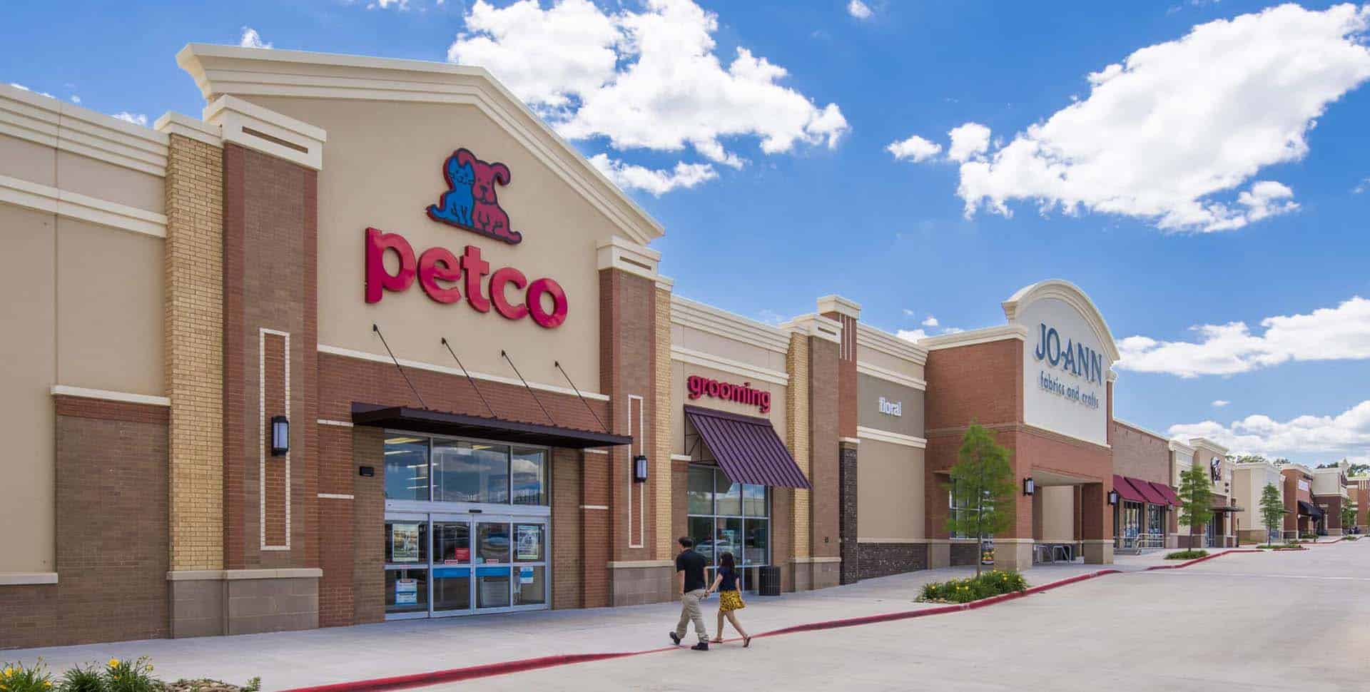 Petco storefront next to Joann Fabrics in a strip mall, with people walking on the sidewalk under a blue sky with clouds.