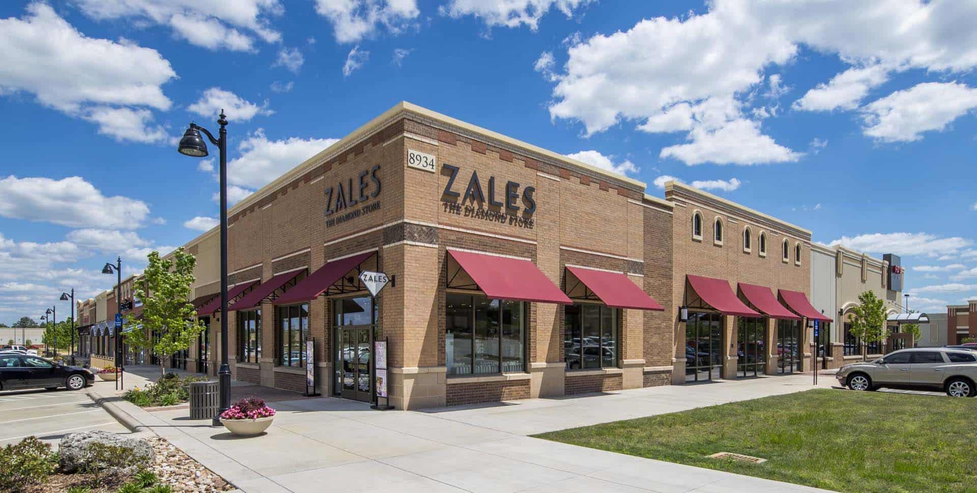 A brick Zales jewelry store with red awnings on a street corner, surrounded by a parking lot and greenery under a blue sky with clouds.