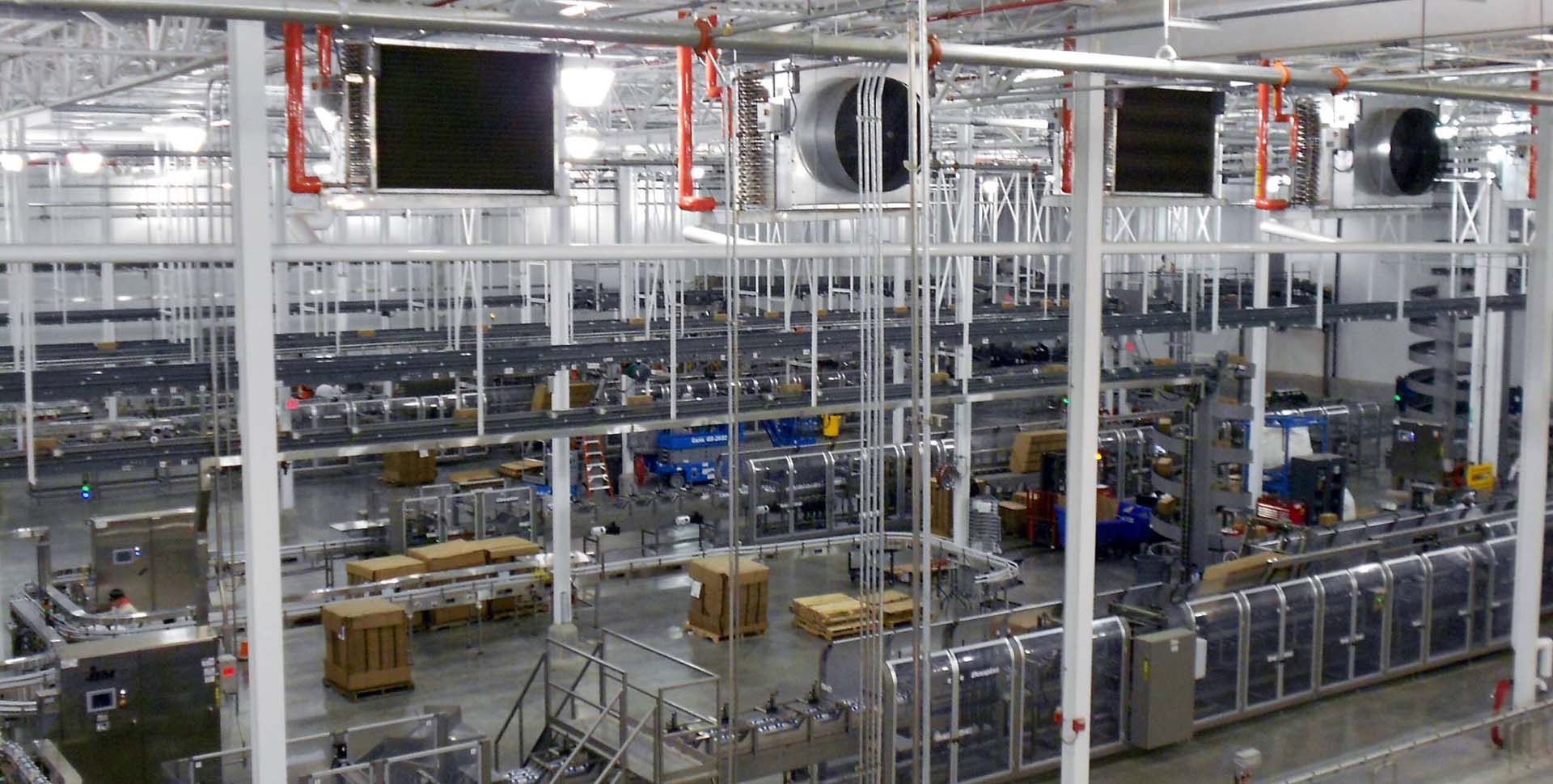 Large industrial factory interior with rows of automated conveyor belts, machinery, and equipment, under bright overhead lighting.