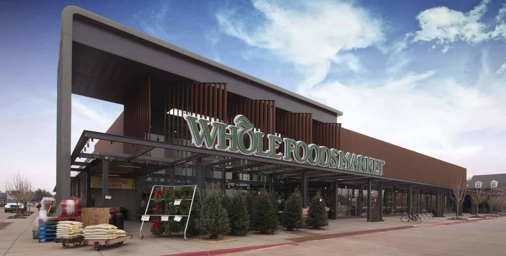 Exterior view of a Whole Foods Market store with potted plants, trees, and bags of goods displayed near the entrance under a partly cloudy sky.