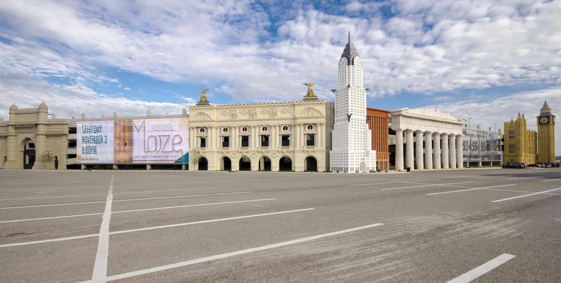 An empty parking lot in front of buildings with various architectural styles, including classical columns and modern skyscraper-like structures, under a partly cloudy sky.
