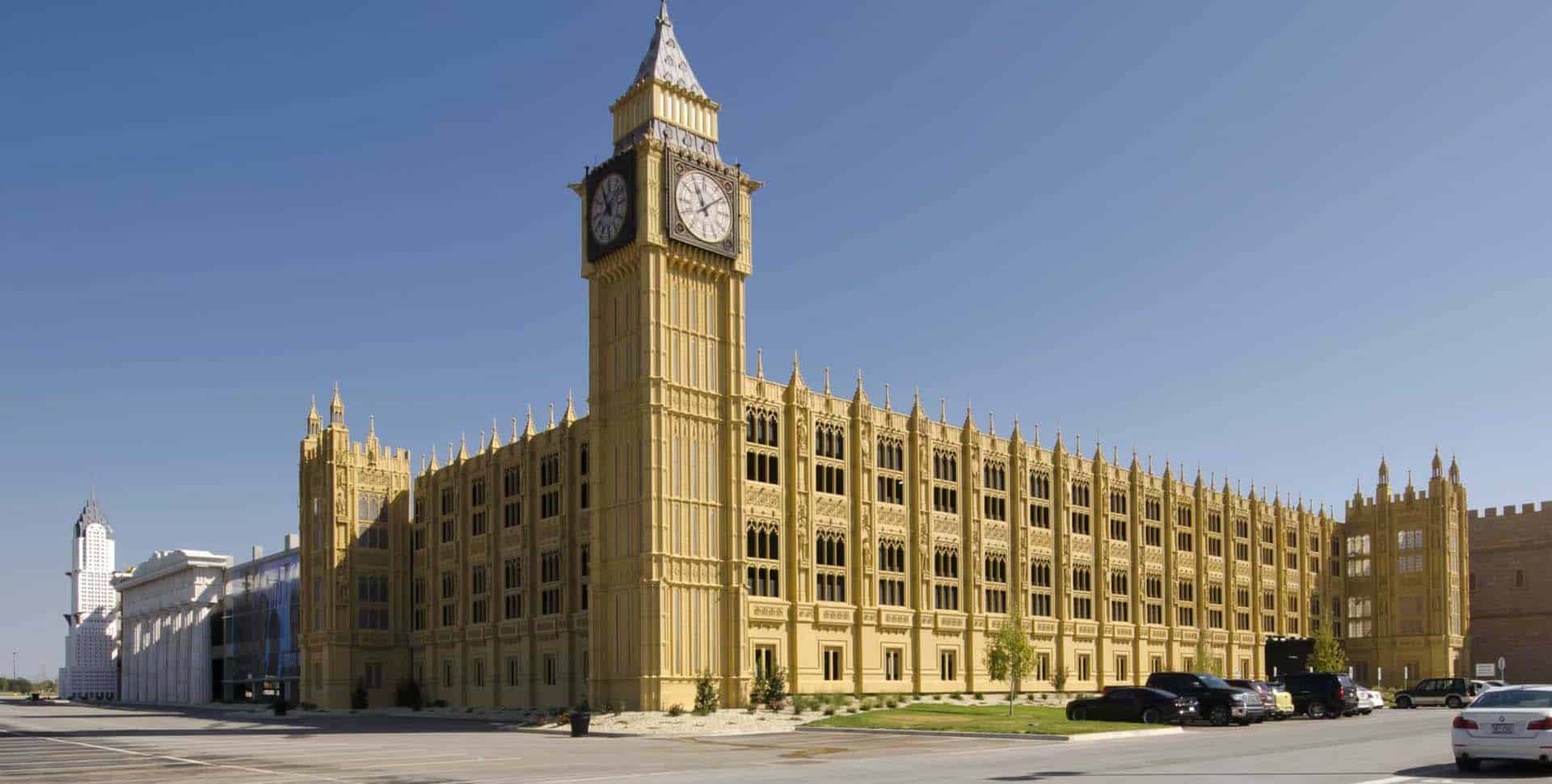 A large building resembling London’s Big Ben and the Palace of Westminster stands in a parking lot under a clear blue sky, with cars parked in front.