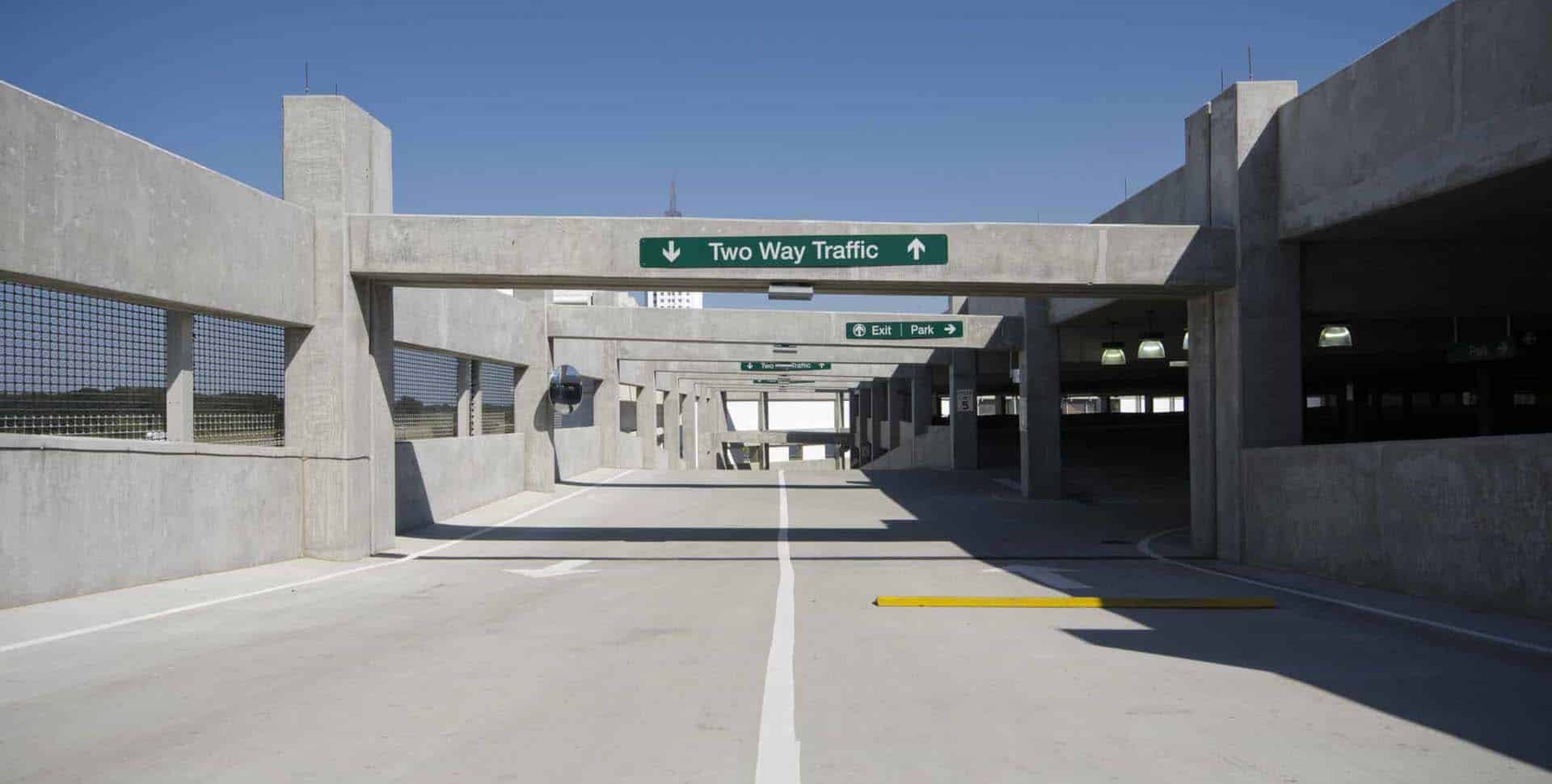 Concrete parking garage with directional signs indicating two-way traffic and sections for exit and parking under clear blue sky.