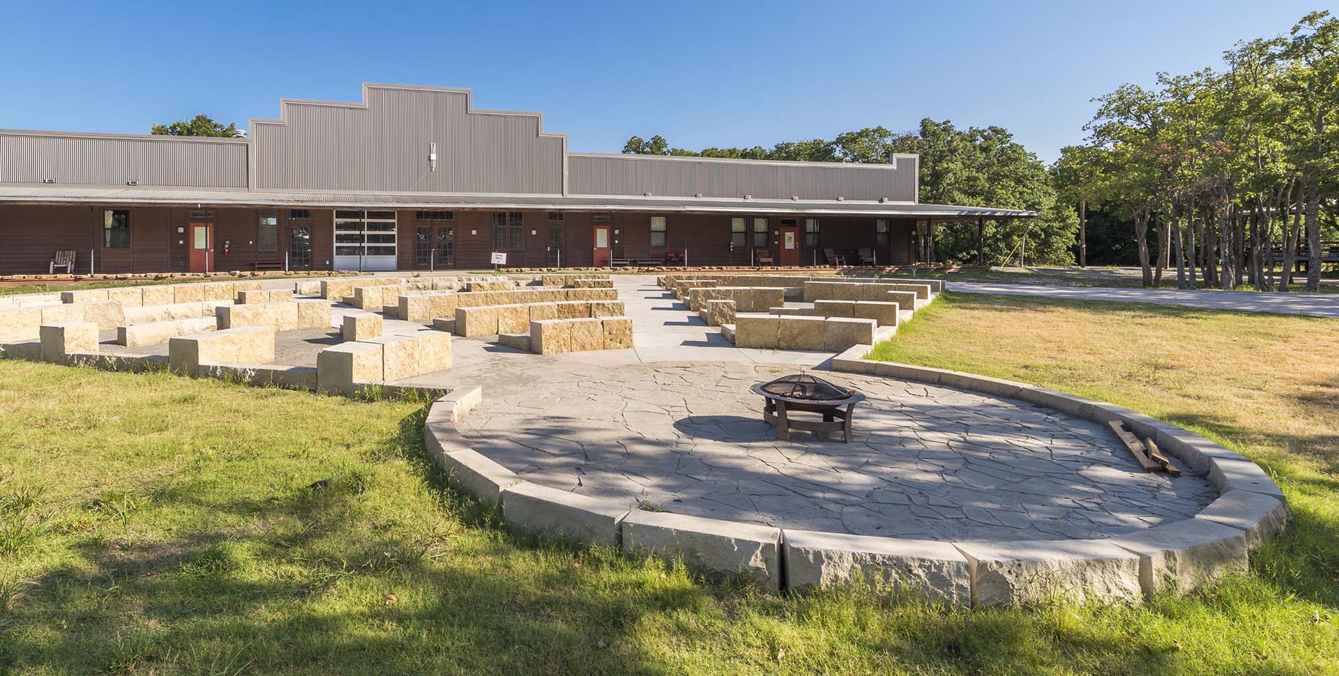 Outdoor amphitheater with stone seating and a central fire pit in front of a large building, surrounded by grass and trees under a clear sky.