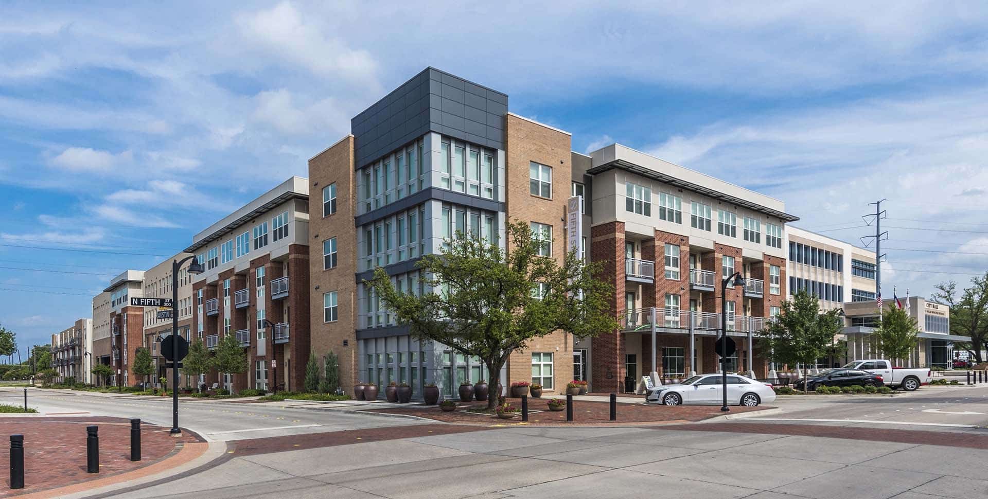 Modern mixed-use building with brick and glass facade on a street corner, featuring balconies, trees, parked cars, and a clear sky overhead.
