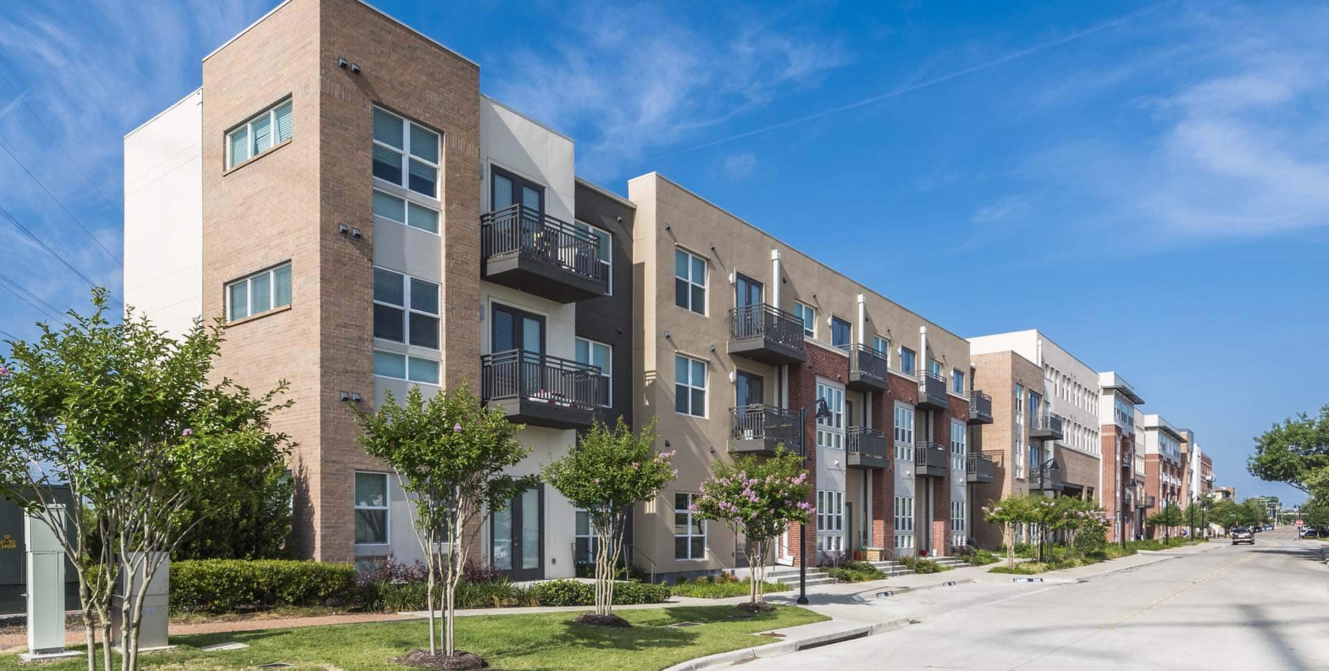 A row of modern, multi-story apartment buildings with balconies and large windows lines a quiet street on a clear, sunny day.