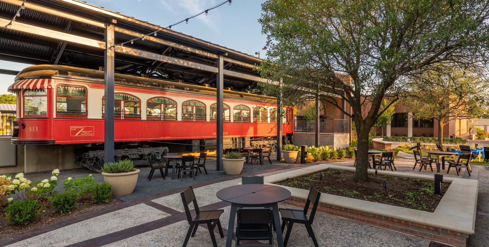 Outdoor seating area with round tables and chairs, large potted plants, and a red vintage streetcar under a covered structure next to a brick building.
