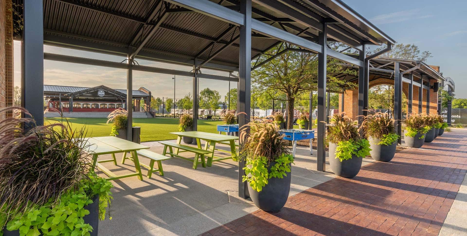 Outdoor pavilion with picnic tables and large potted plants beside a grassy lawn, under a metal roof, on a sunny day.