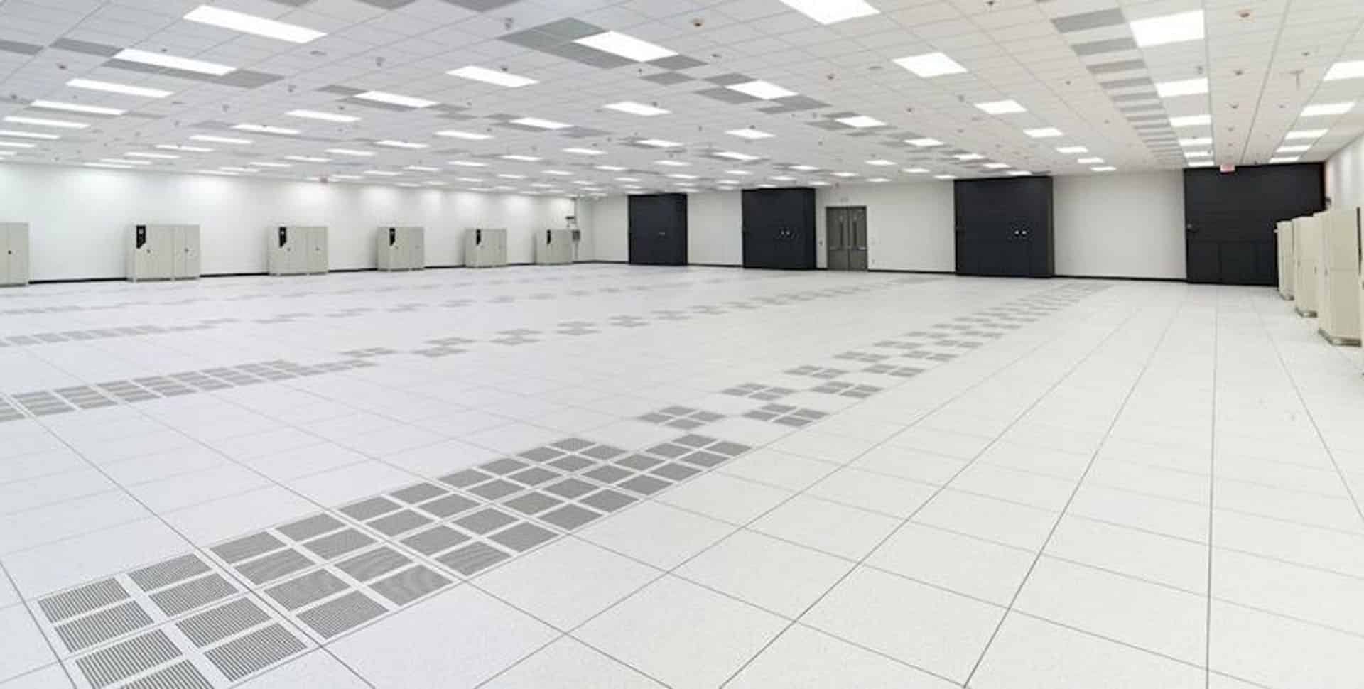 Large, empty data center room with white floor tiles, ceiling lights, rows of ventilation grids, and several server cabinets along white walls.