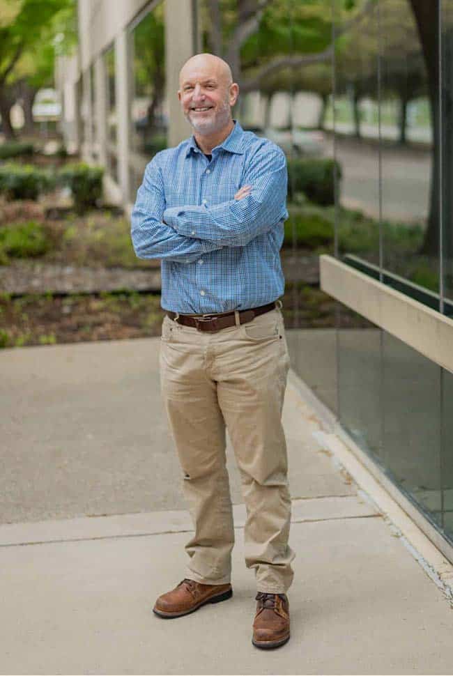 A man with a bald head and gray beard stands outside near a building, wearing a blue checked shirt, khaki pants, and brown shoes. His arms are crossed and he is smiling.