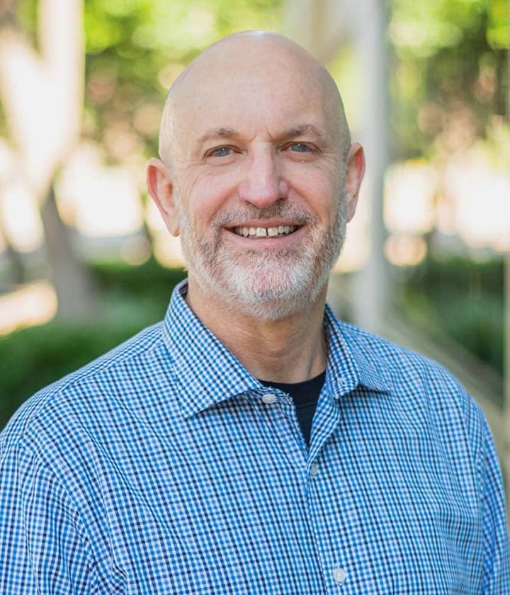A middle-aged man with a bald head and gray beard is smiling, wearing a blue checkered shirt, standing outdoors with greenery and sunlight in the background.