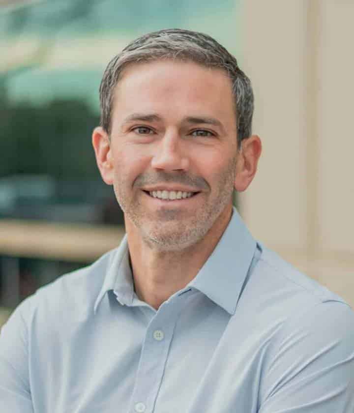 A man with short gray hair and a trimmed beard is smiling, wearing a light blue collared shirt, standing outdoors in front of a blurred background.