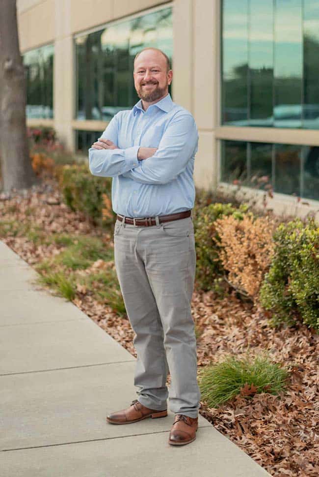 A man with a beard stands on a sidewalk outside a building with large windows, arms crossed, wearing a light blue shirt, gray pants, and brown shoes.