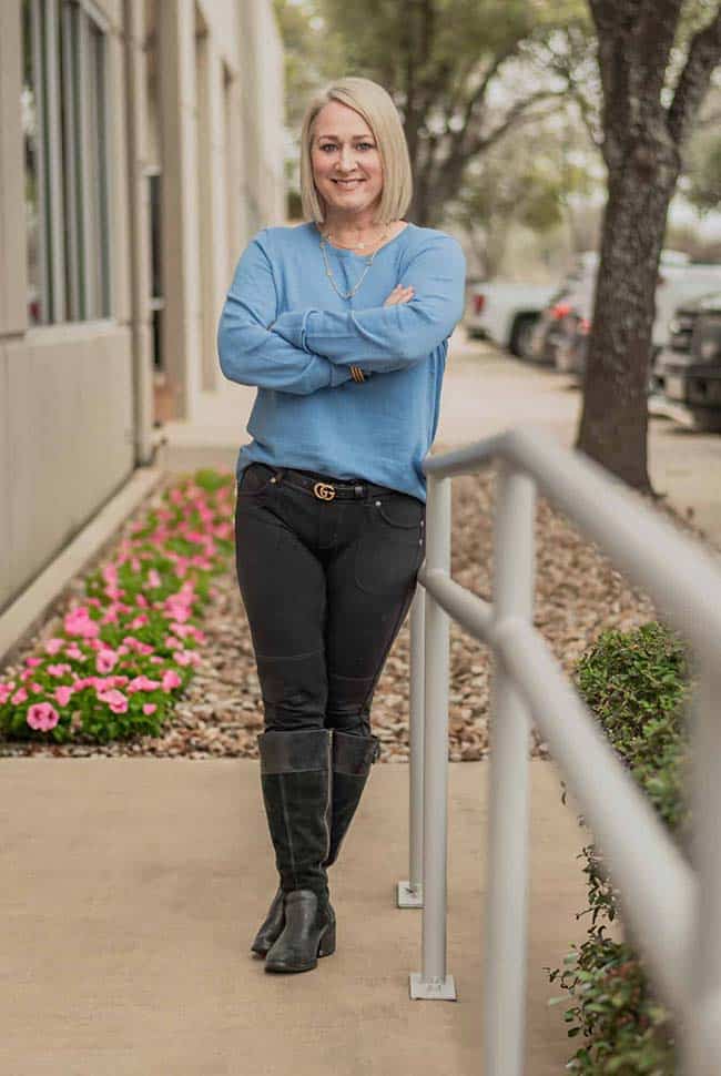 A woman with blonde hair, wearing a blue sweater, black pants, and black boots, stands with arms crossed on a sidewalk near a building and flower bed.