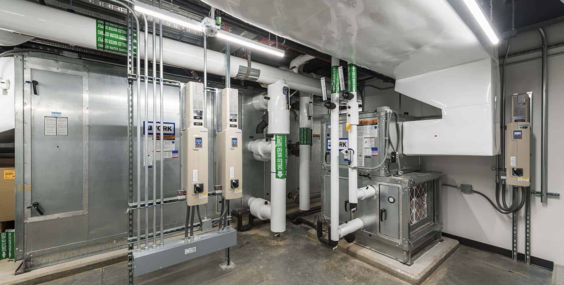 Room with industrial HVAC equipment, including ductwork, control panels, insulated pipes labeled "chilled water supply" and "hot water supply," and various electrical components.