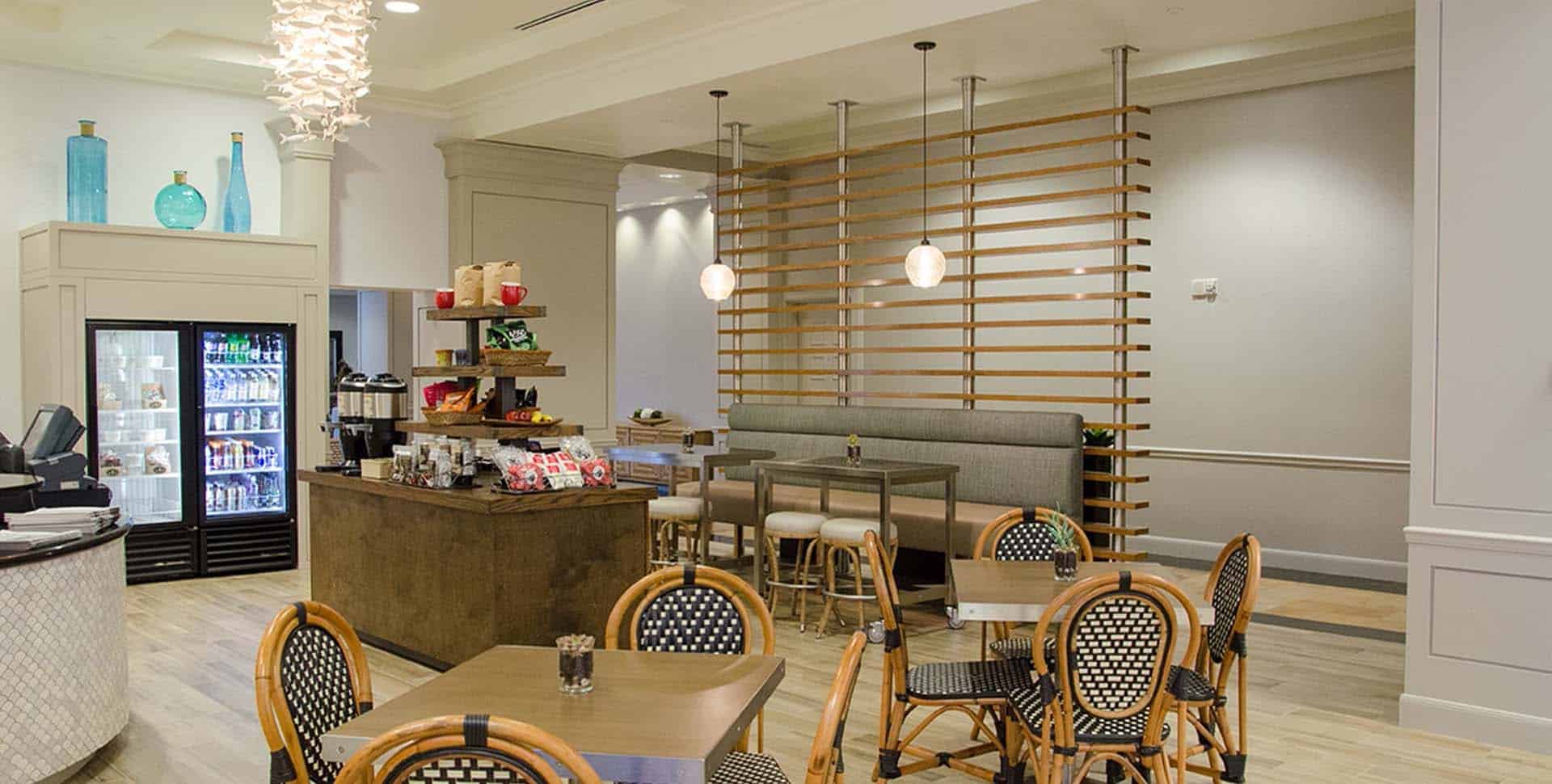 Modern café interior with round tables, woven chairs, a refrigerated display, a coffee station, and a booth with pendant lights and wooden room divider.