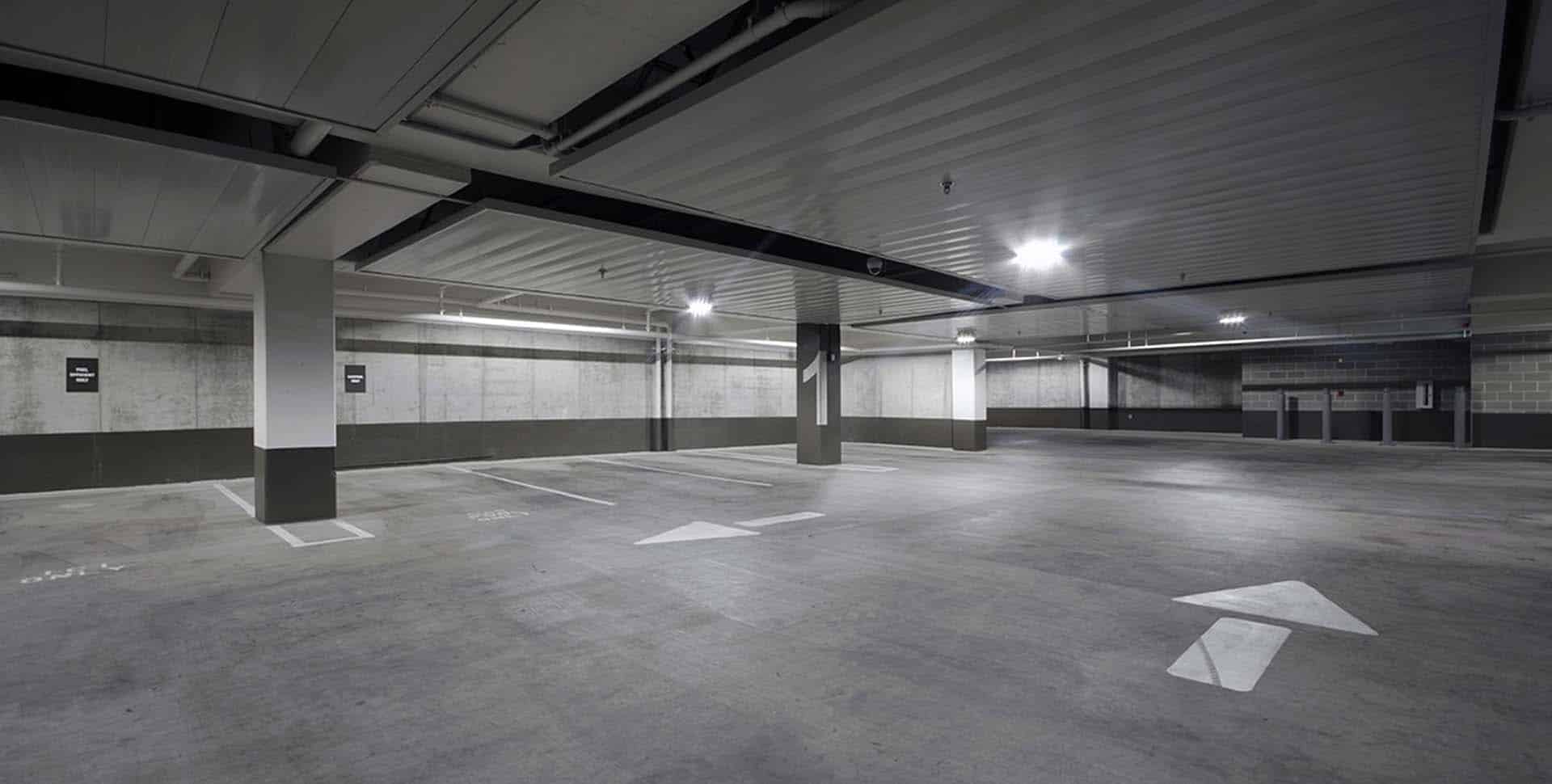 Empty underground parking garage with concrete floors, support columns, overhead lighting, and directional arrows painted on the ground.