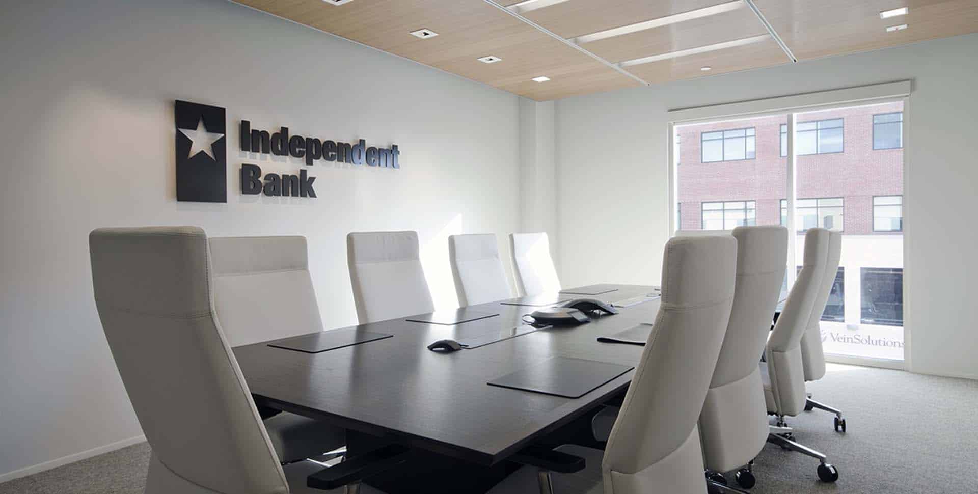 Modern conference room with a long table, eight white chairs, conference phone, and "Independent Bank" logo on the wall near a large window.