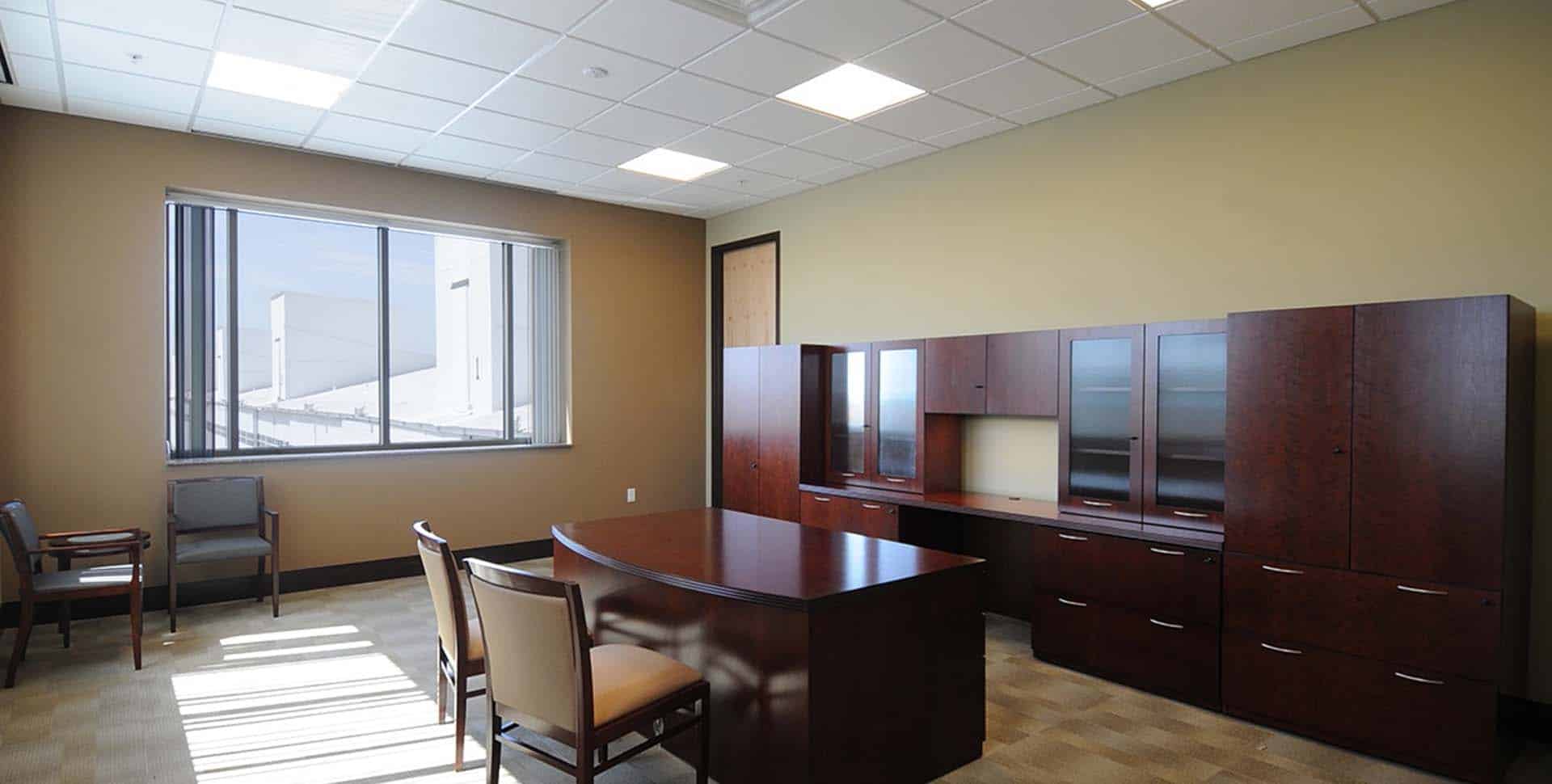 A modern office with a large wooden desk, chairs, cabinets, and a window letting in natural light.
