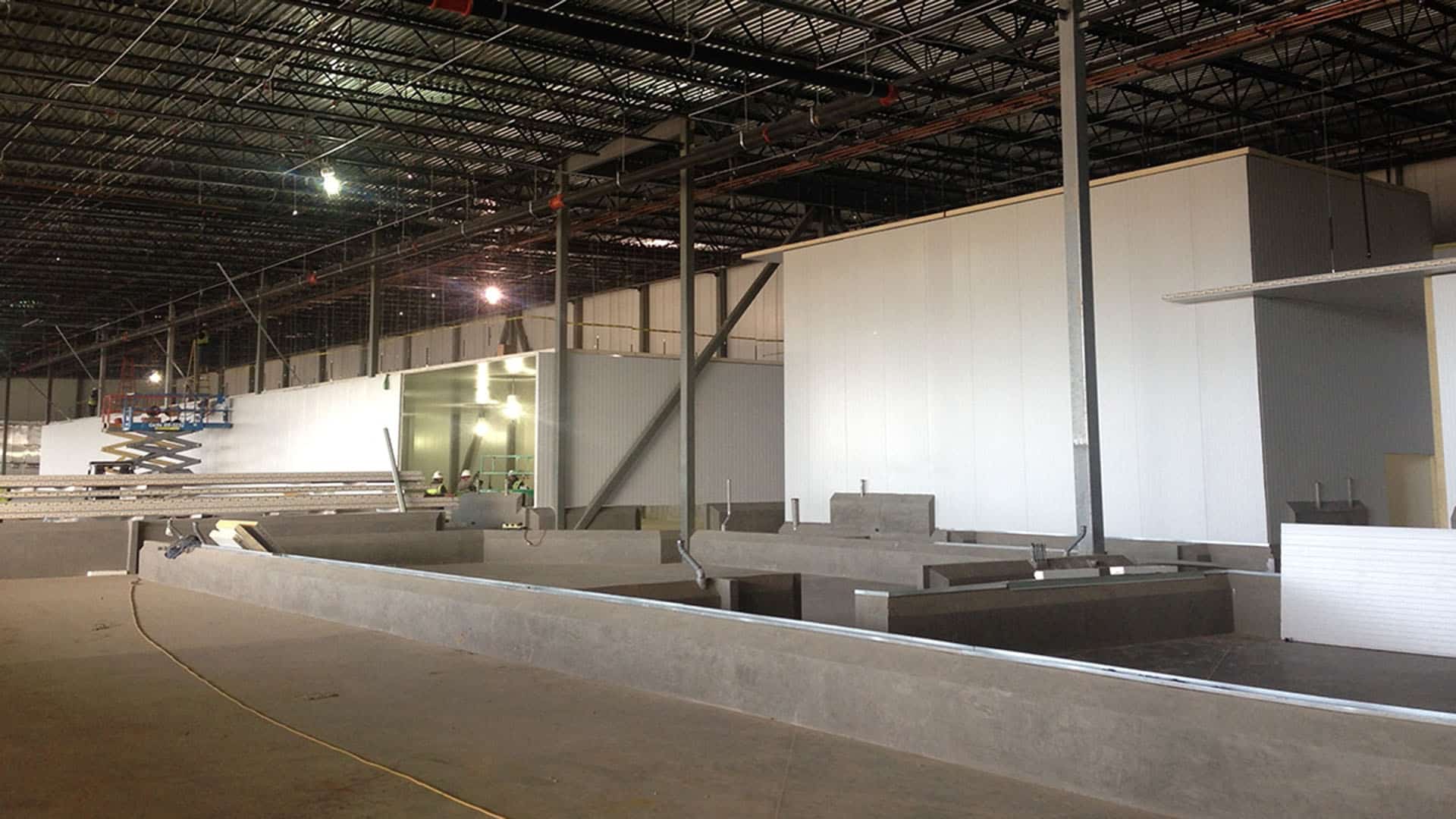 Large industrial warehouse with unfinished walls, exposed ceiling beams, construction materials, and workers using a scissor lift.