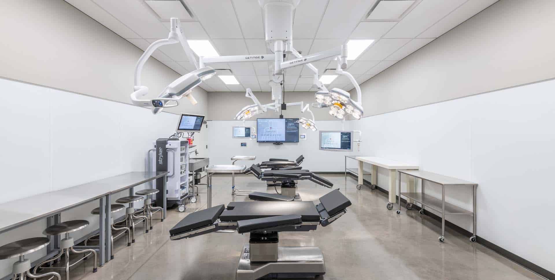 A modern, sterile operating room with surgical tables, overhead lights, multiple monitors, medical equipment, and stools along one wall.