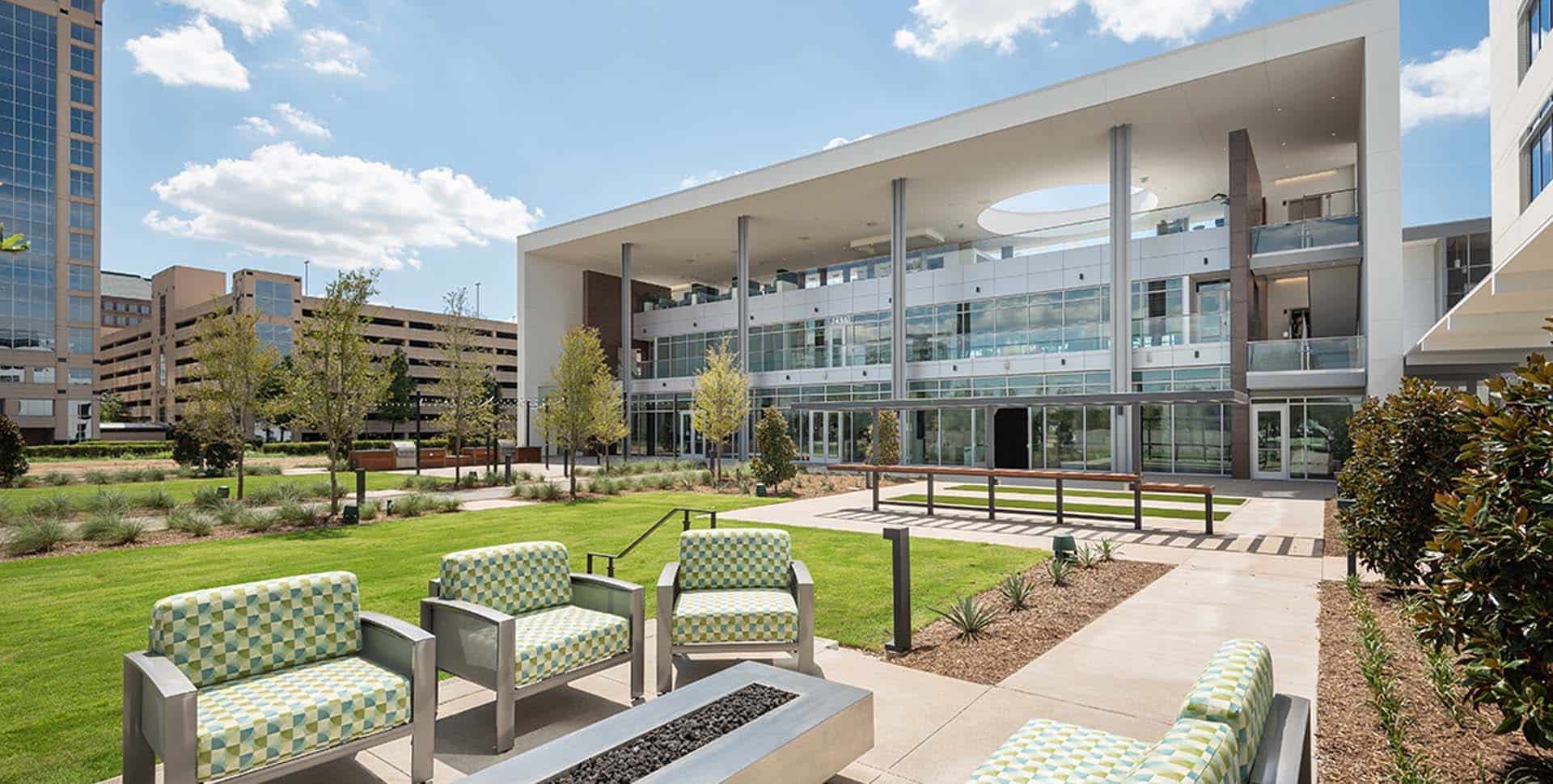 Modern office building with large windows and an outdoor seating area featuring patterned chairs and a fire pit, surrounded by landscaped greenery under a partly cloudy sky.