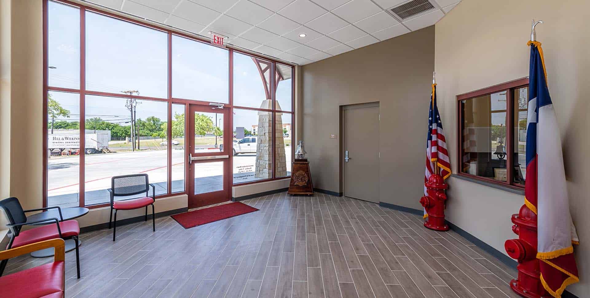 A lobby with large windows, chairs, and two flags beside a door. Fire hydrant decor and a window to an adjacent office are also visible.