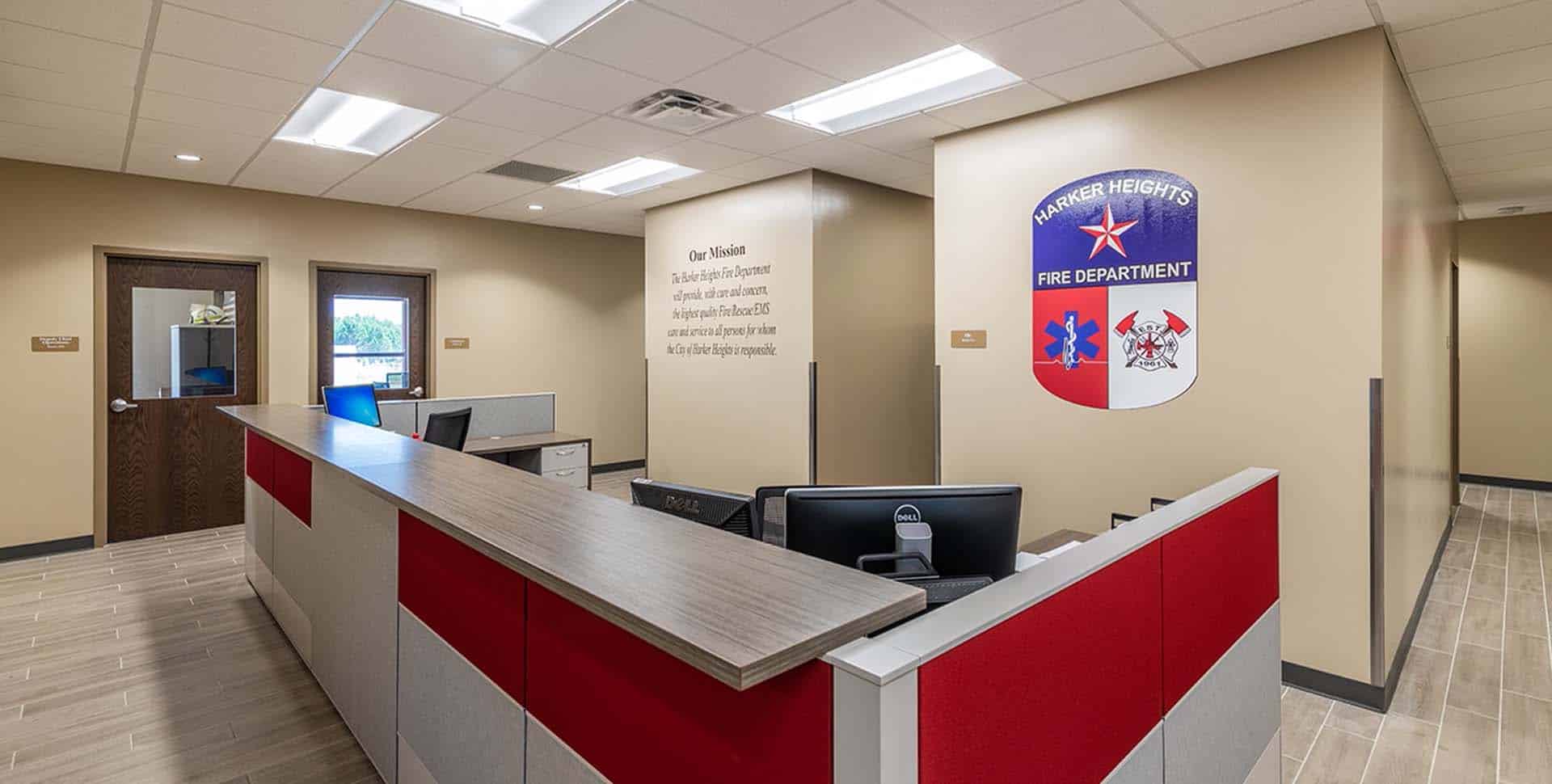 Reception area of a fire department office with workstations, beige walls, a mission statement, and the fire department emblem on the wall.