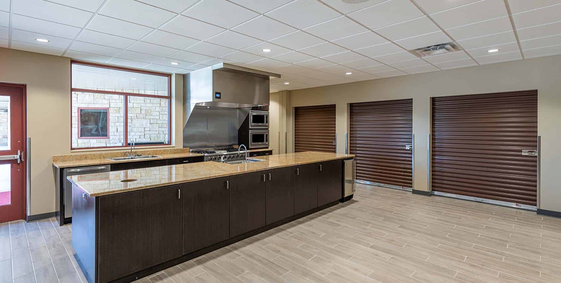 Modern commercial kitchen with a large island, stainless steel appliances, wood cabinets, tile flooring, and three brown roll-up doors along the back wall.