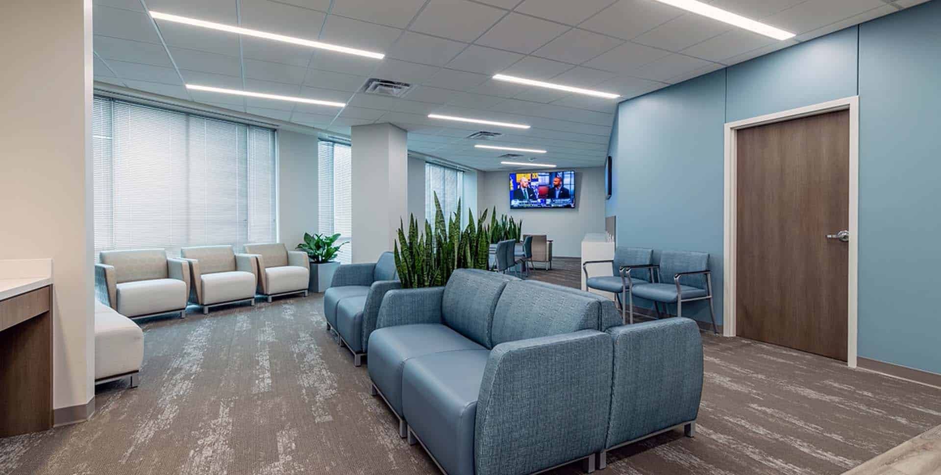 Modern waiting room with blue and beige chairs, a row of plants, a TV on the wall, and large windows letting in natural light.