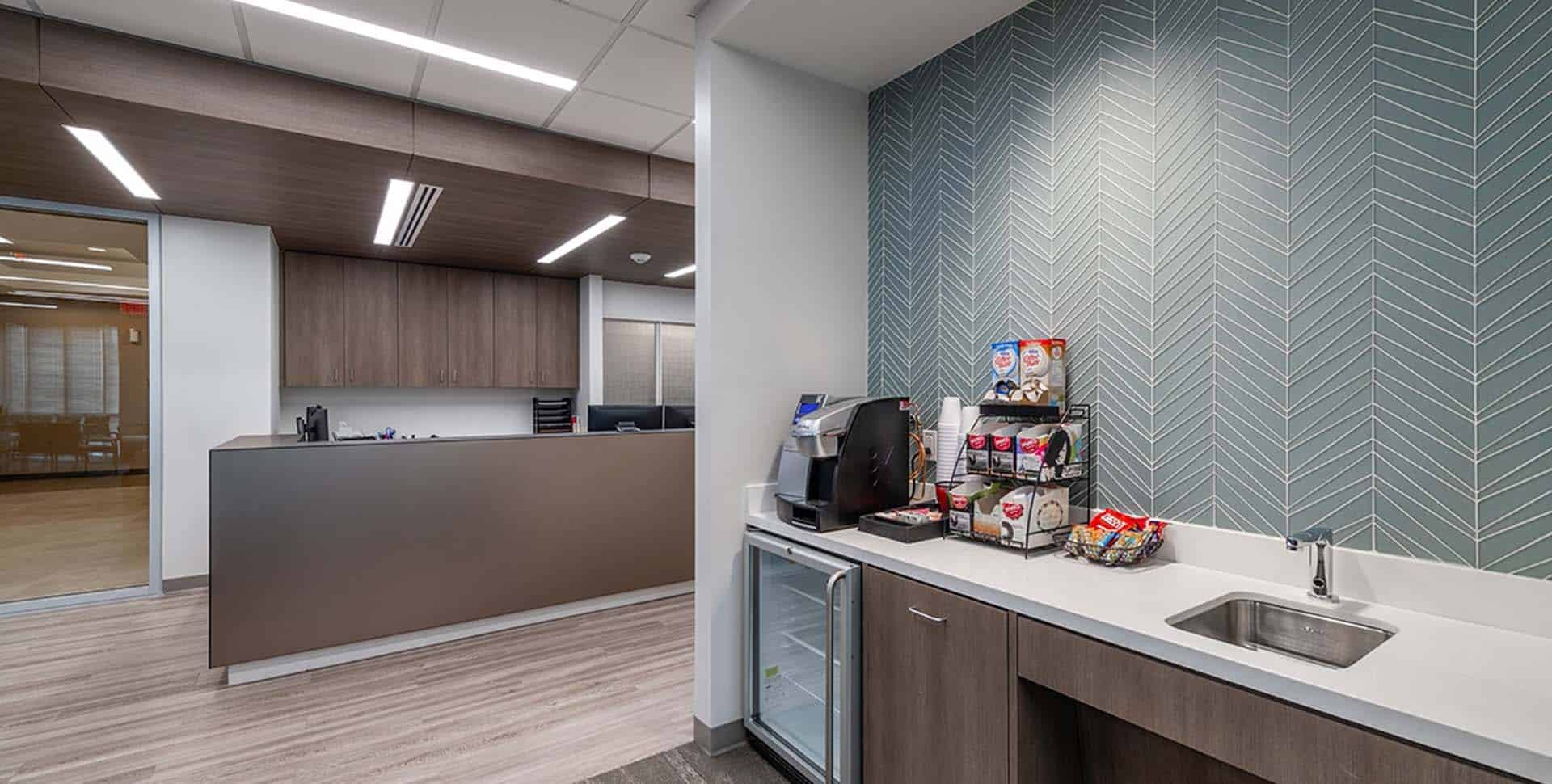 Modern office break room with a coffee station, snack display, small refrigerator, sink, and geometric patterned wall; open workspace visible in the background.