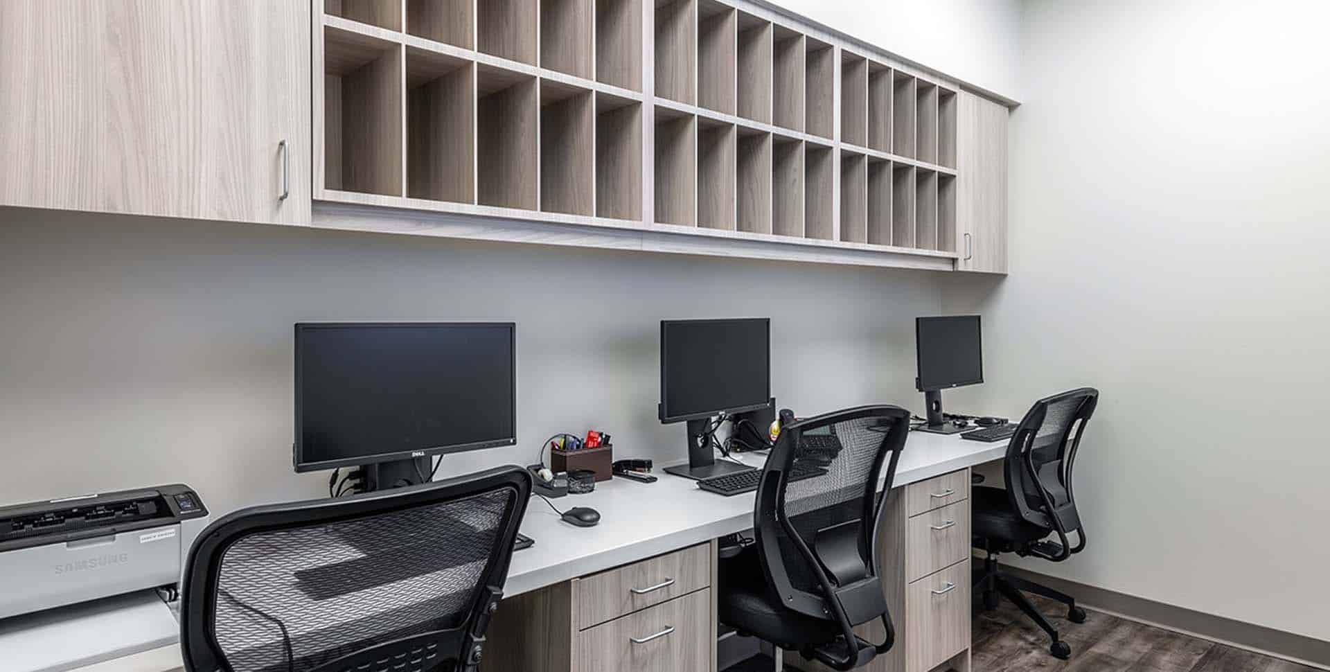 A modern office workspace with three computer desks, black chairs, monitors, a printer, and wall-mounted shelves with open cubbies above.