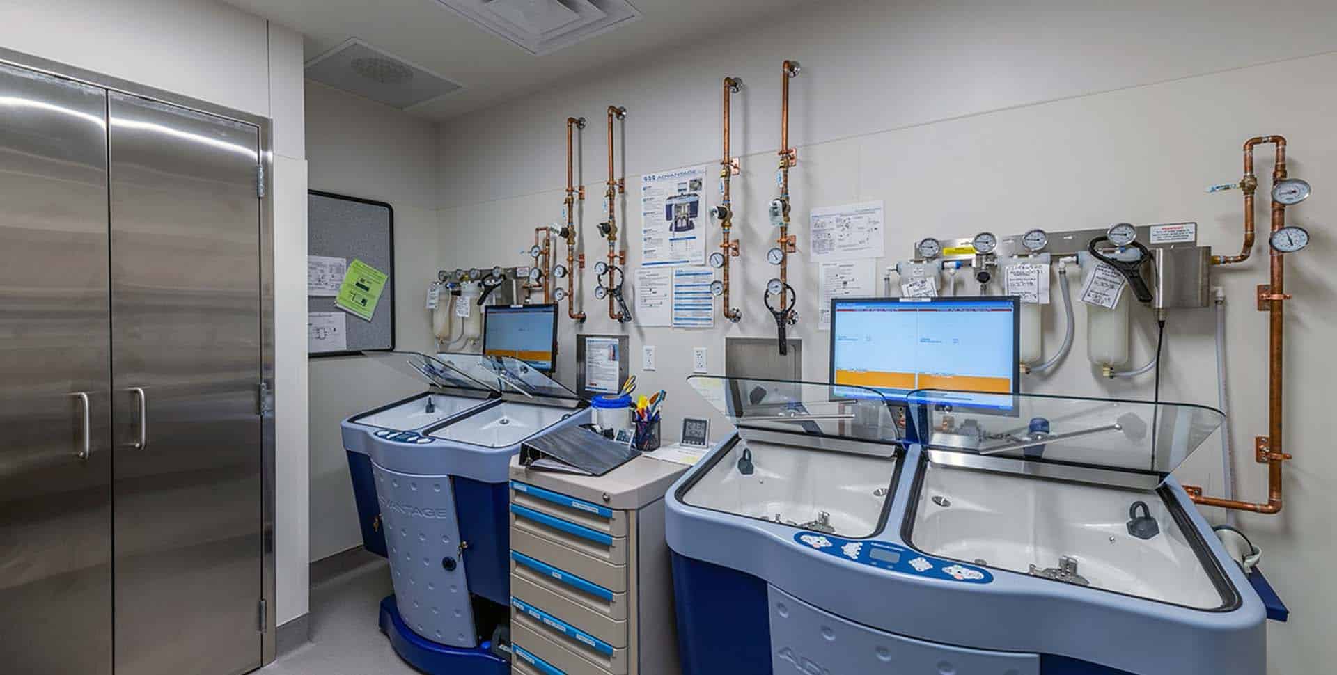 A laboratory room with two blue and silver workstations, monitors, medical equipment, wall-mounted gauges, copper pipes, and a stainless steel cabinet.