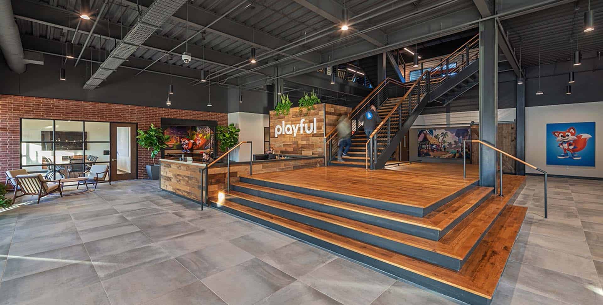Modern office lobby with wooden stairs, "playful" sign, glass-walled meeting room, seating area, large indoor plants, and colorful artwork on the walls.