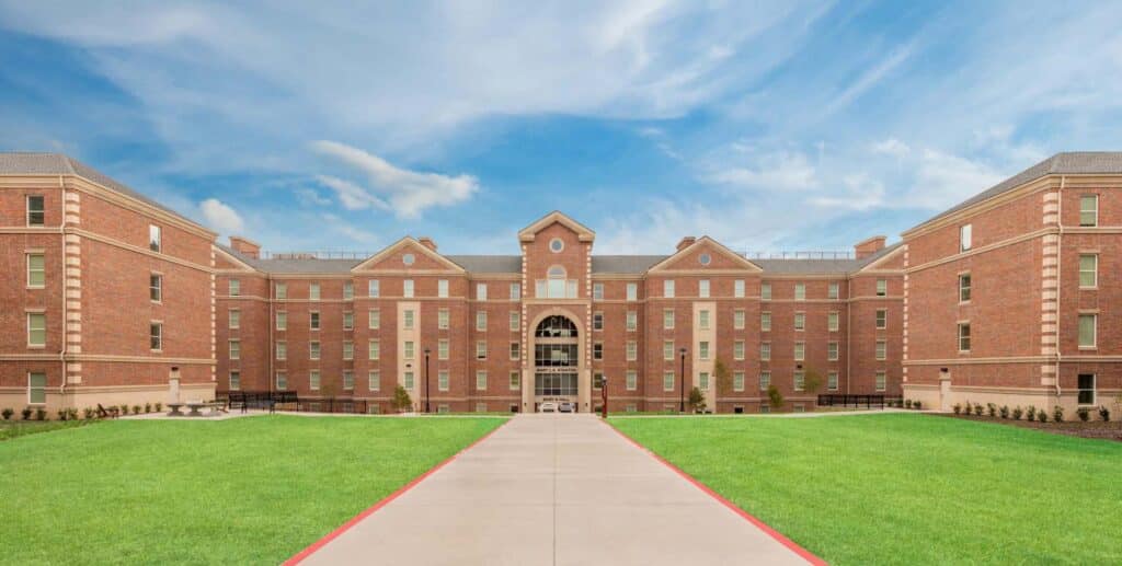 Large brick dormitory building with symmetrical wings, multiple windows, and a central entrance, set against a blue sky and bordered by a wide sidewalk and green lawn.