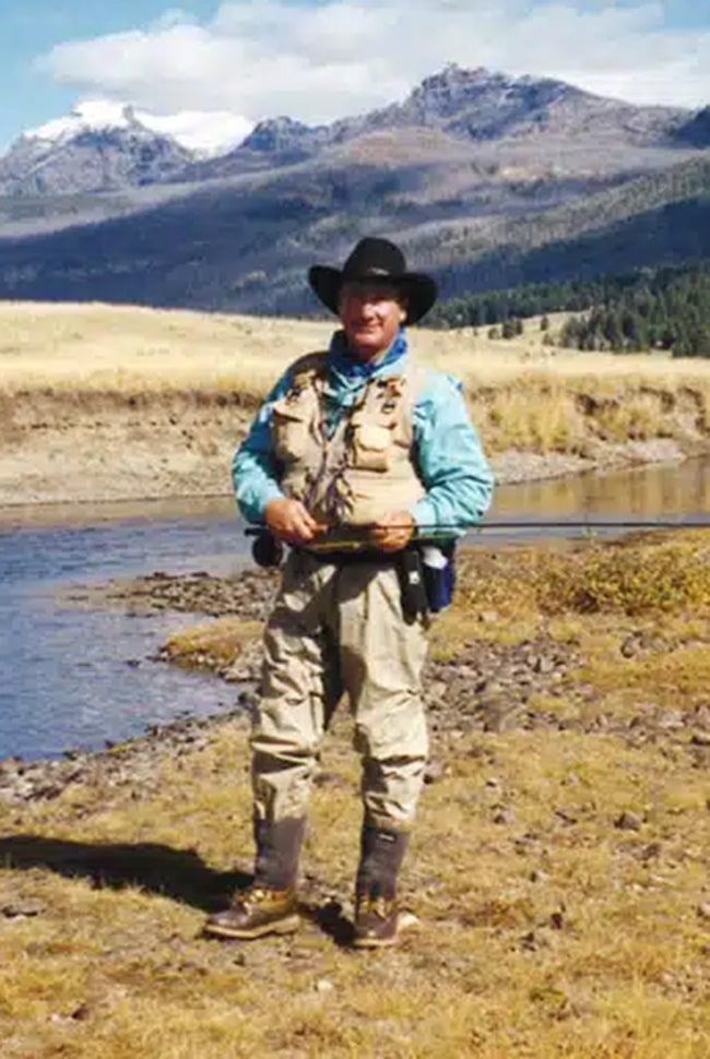 A person in fishing gear stands by the edge of a river with mountains and grassy fields in the background.