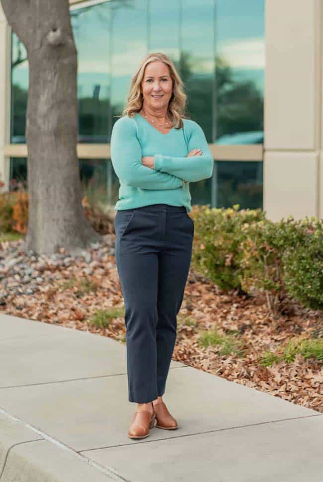 A woman with blonde hair stands on a sidewalk with arms crossed, wearing a light blue sweater, dark pants, and brown shoes, in front of a building and landscaped area.